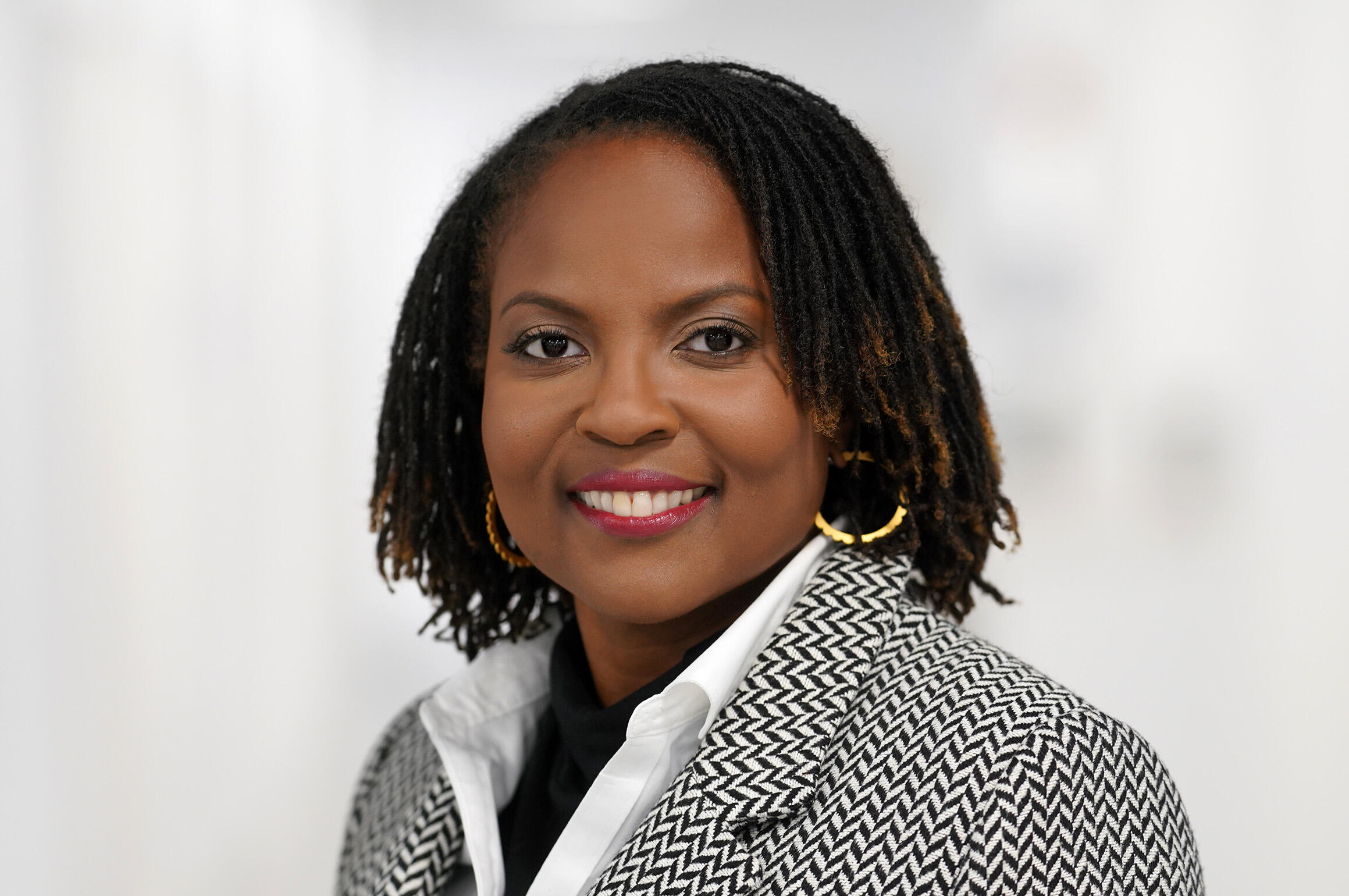 A Black Woman wearing a black and white sports coat, a white button up shirt, and gold hoop earrings. 
