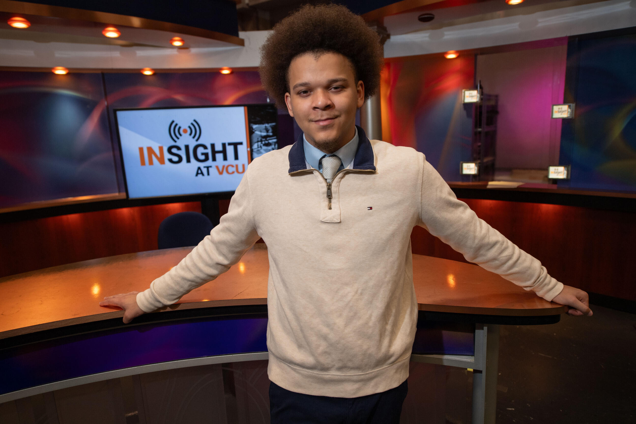 A photo of a man standing inside a TV studio set. Behind him is a screen that says \"INSIGHT AT VCU\" in black and yellow letters. 