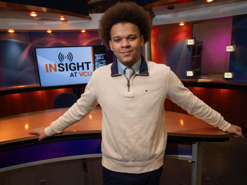 A photo of a man standing inside a TV studio set. Behind him is a screen that says \"INSIGHT AT VCU\" in black and yellow letters. 