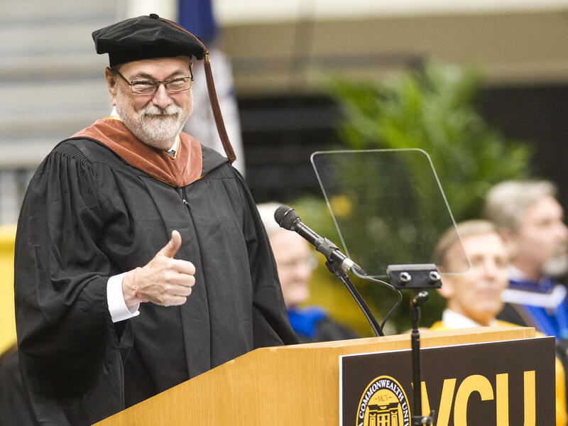 A photo of a man wearing a doctorate cap and gown giving a thumbs up at a podium. 