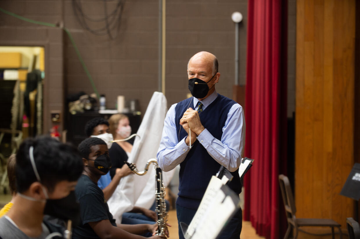 a symphony conductor, speaks to his musicians during a rehearsal. everyone is wearing a mask
