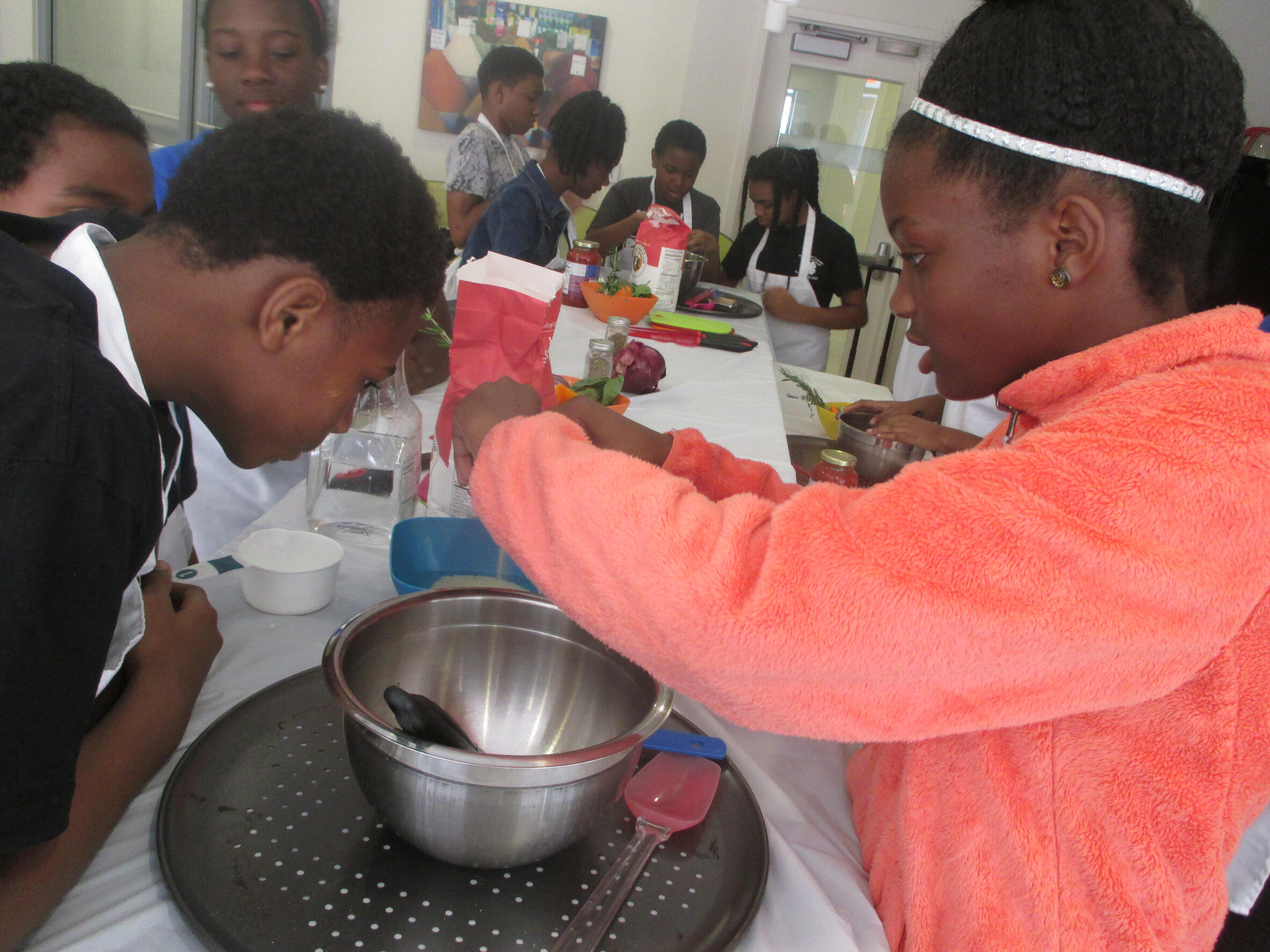 Two youth looking into a mixing bowl, the girl on the right is cracking an egg over it. 