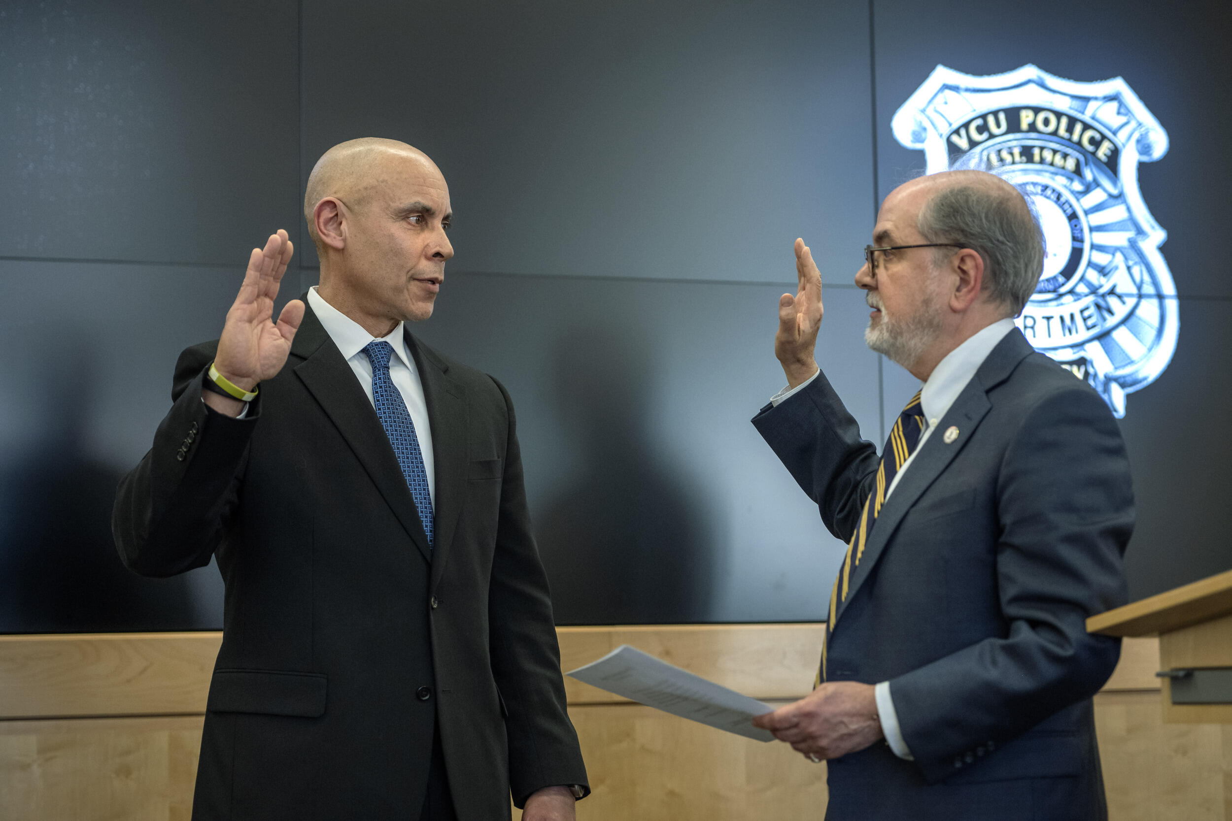 A photo of two men facing each other, both with one arm bent at the elbow with their hand up in the air. The man on the right is holding a stack of papers in his other hand. Both men are wearing suit and ties. 