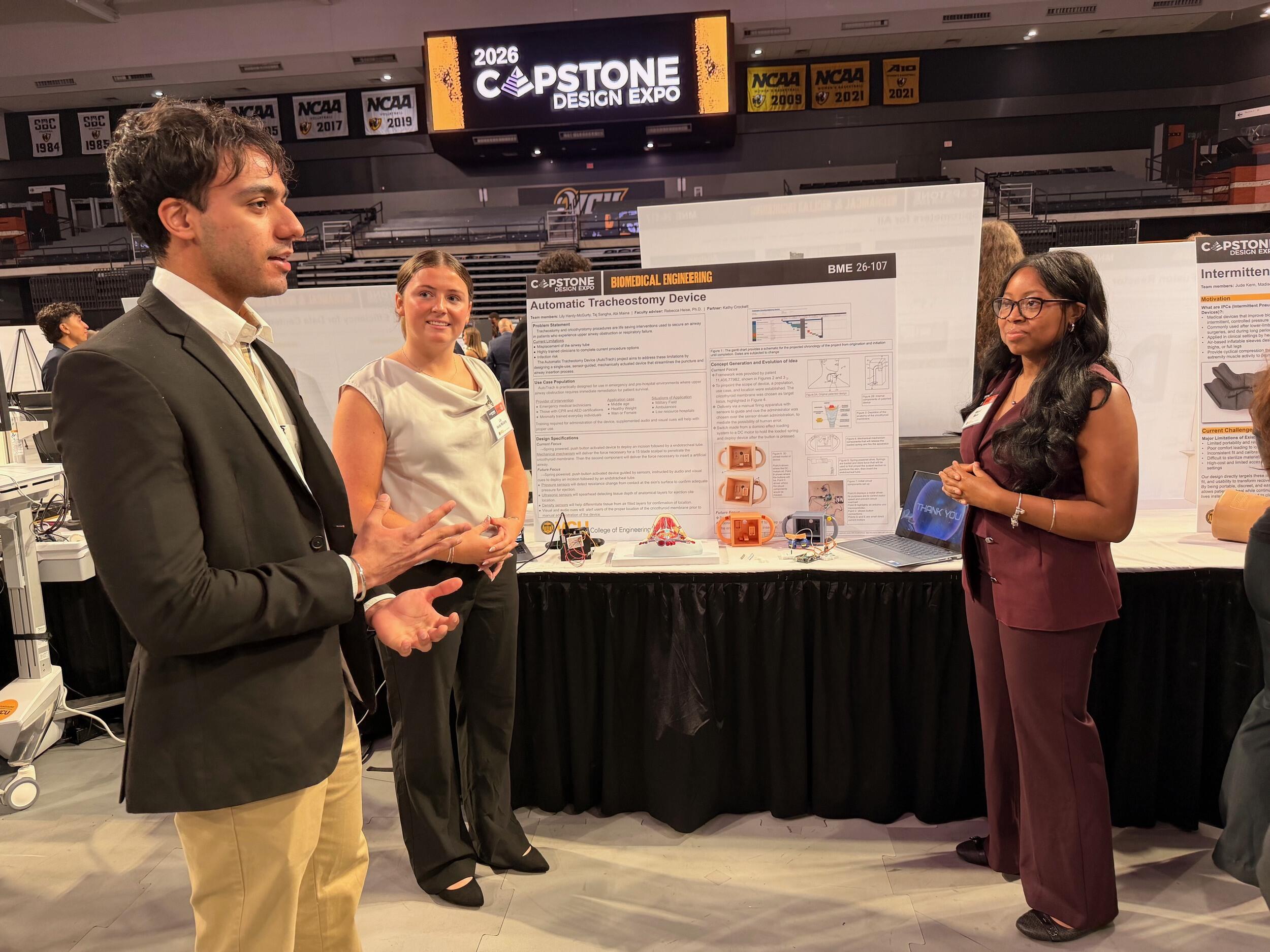 A photo of three people standing around a table with a research poster on it. 