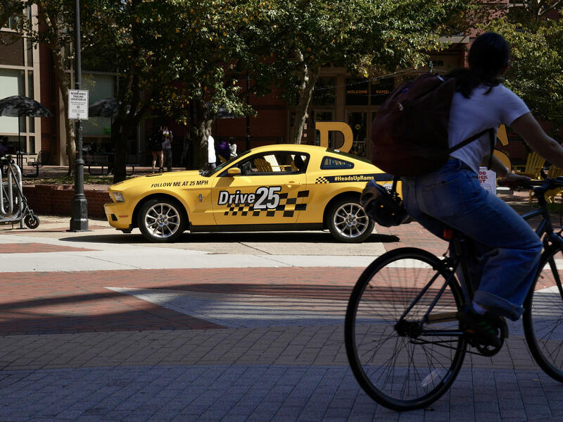 A photo of a yellow sports car that says \"DRIVE 25\" on the side of it. There is a person riding their bike past the car. 