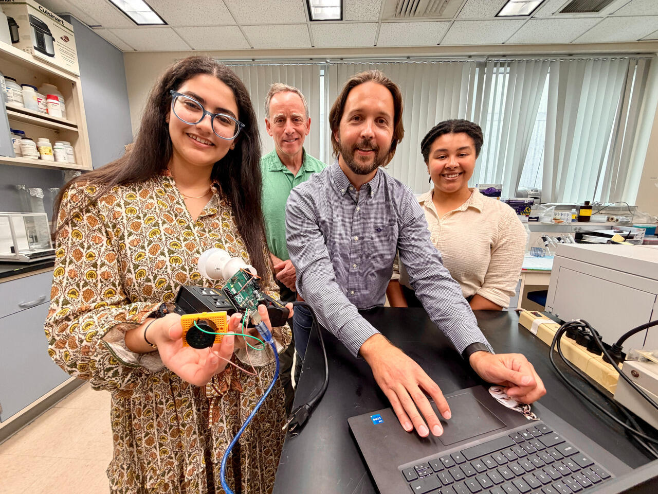 A group photo of four people. The woman on the far left is holding a piece of technology. The man in the center has his hands on an open laptop. Everyone is smiling.