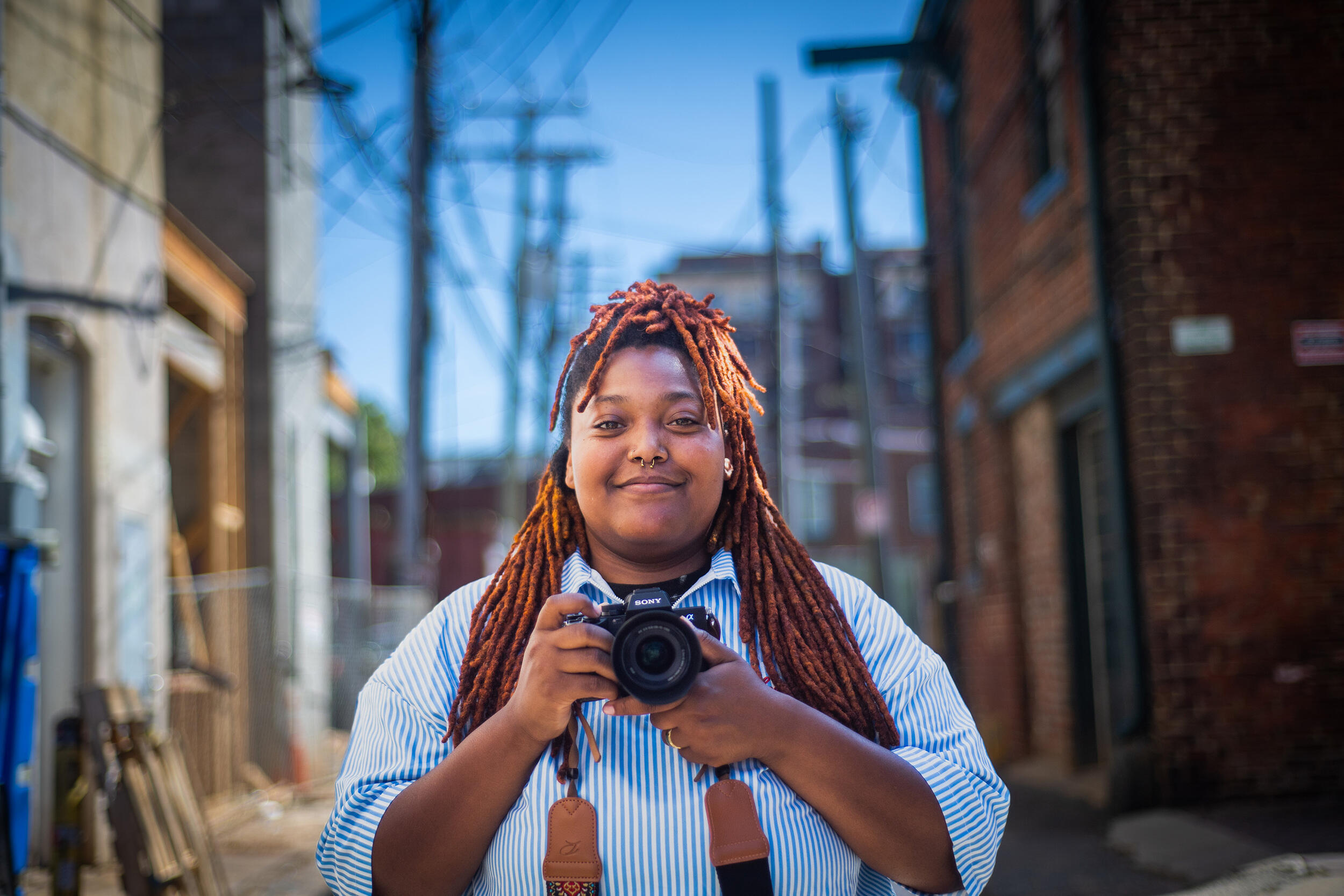A photo of a woman from the chest up. She is standing in an Alley and holding a camera. 