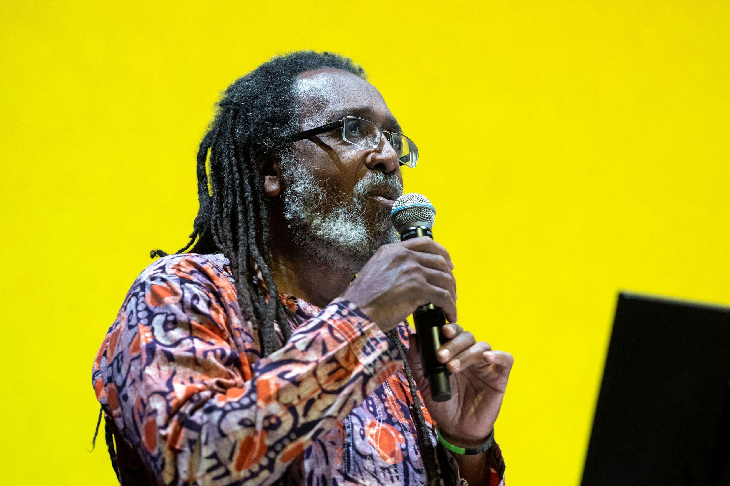 A man with long dreads speaking into a microphone in front of a yellow background. 