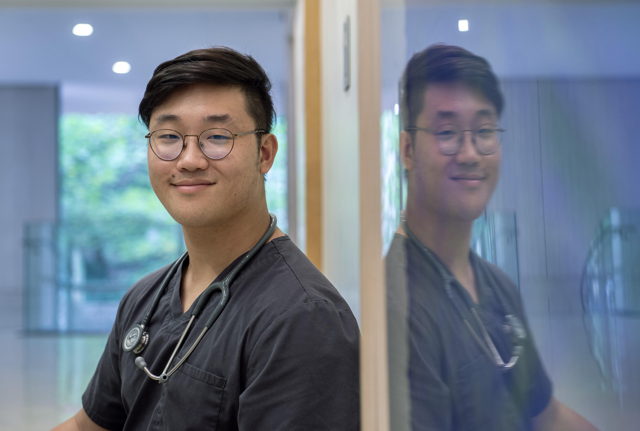 A photo of a man from the chest up standing next to a wall with glass on it. The glass is reflecting the image of the man on it. The man is wearing a medical scrub shirt and a stethoscope around his neck. 