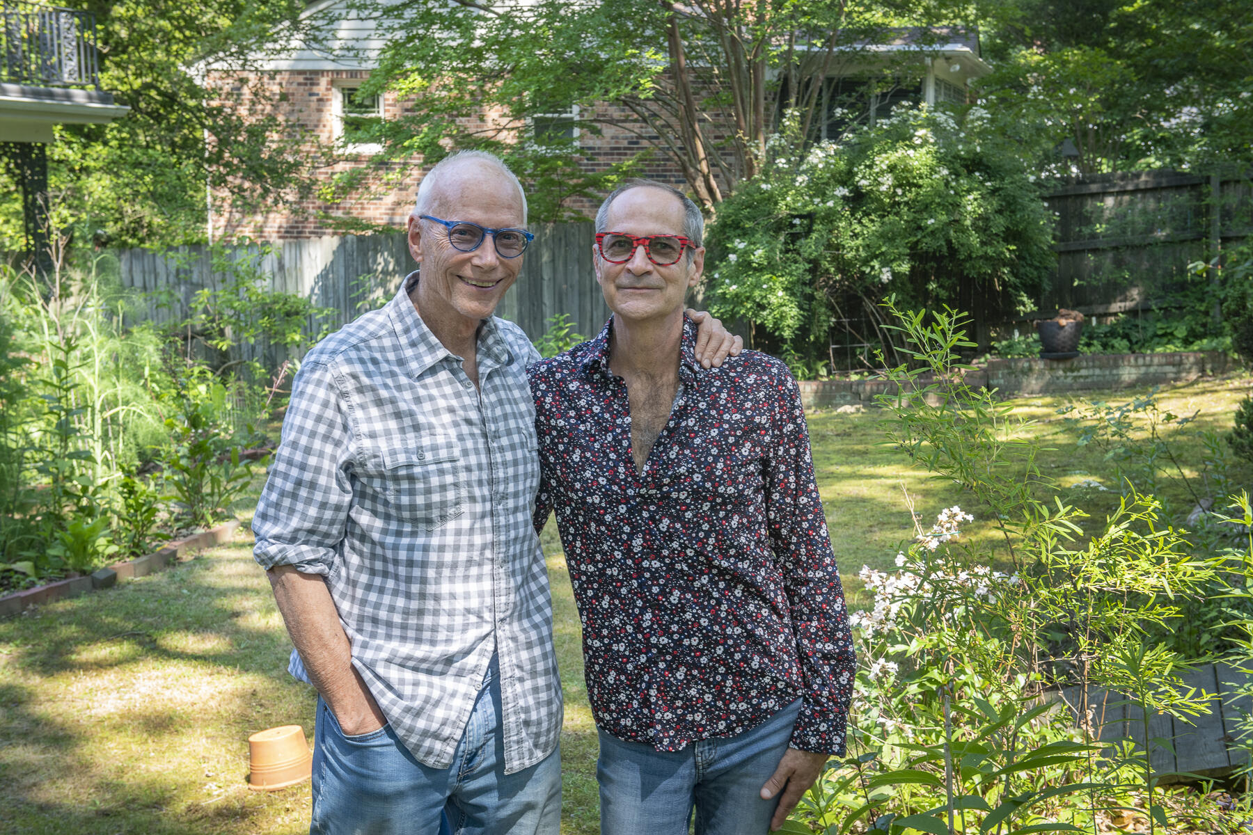 Two men standing next to each other in a garden with one of their arms around each other.