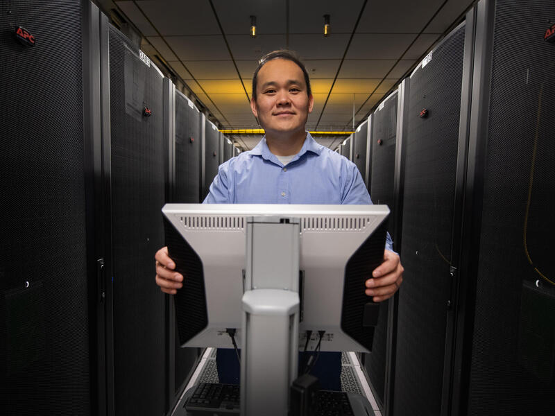 A photo of a man standing behind a computer monitor. 