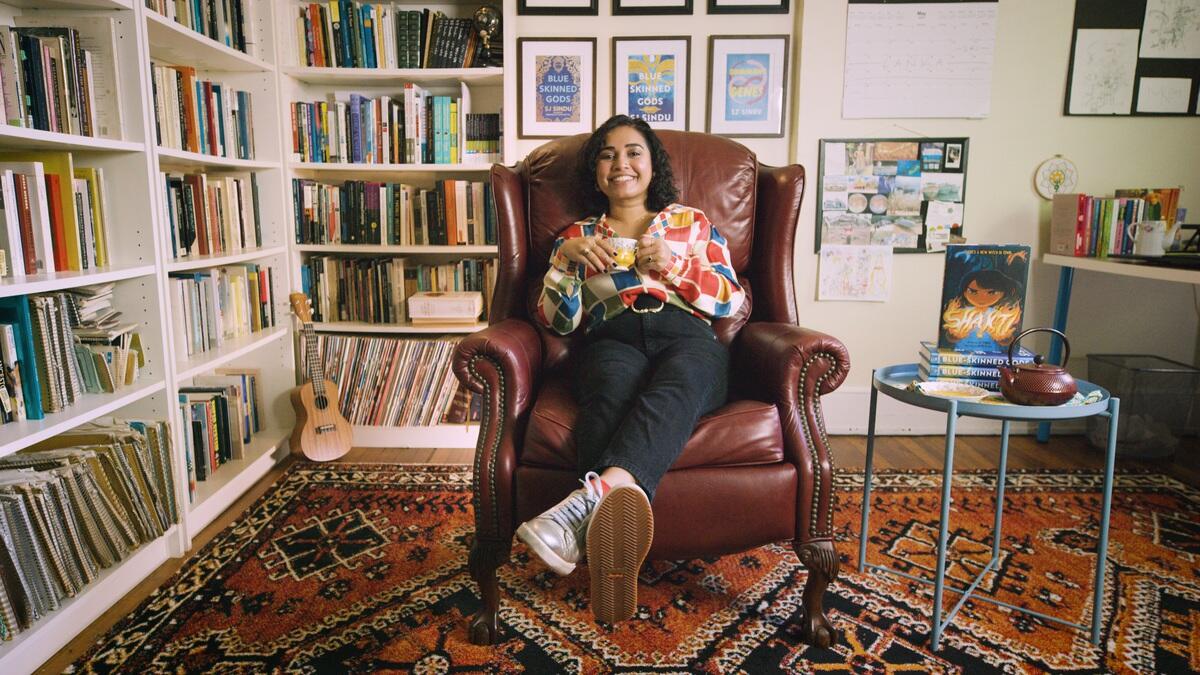 SJ Sindu lounges in a chair holding a cup of tea in an office with bookshelves.