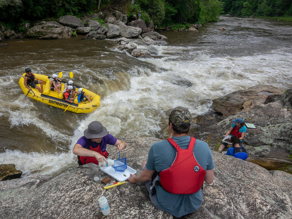 Four researchers conduct research on the shore of a river. A raft with five occupants traverses rapids on the river in the middle background.