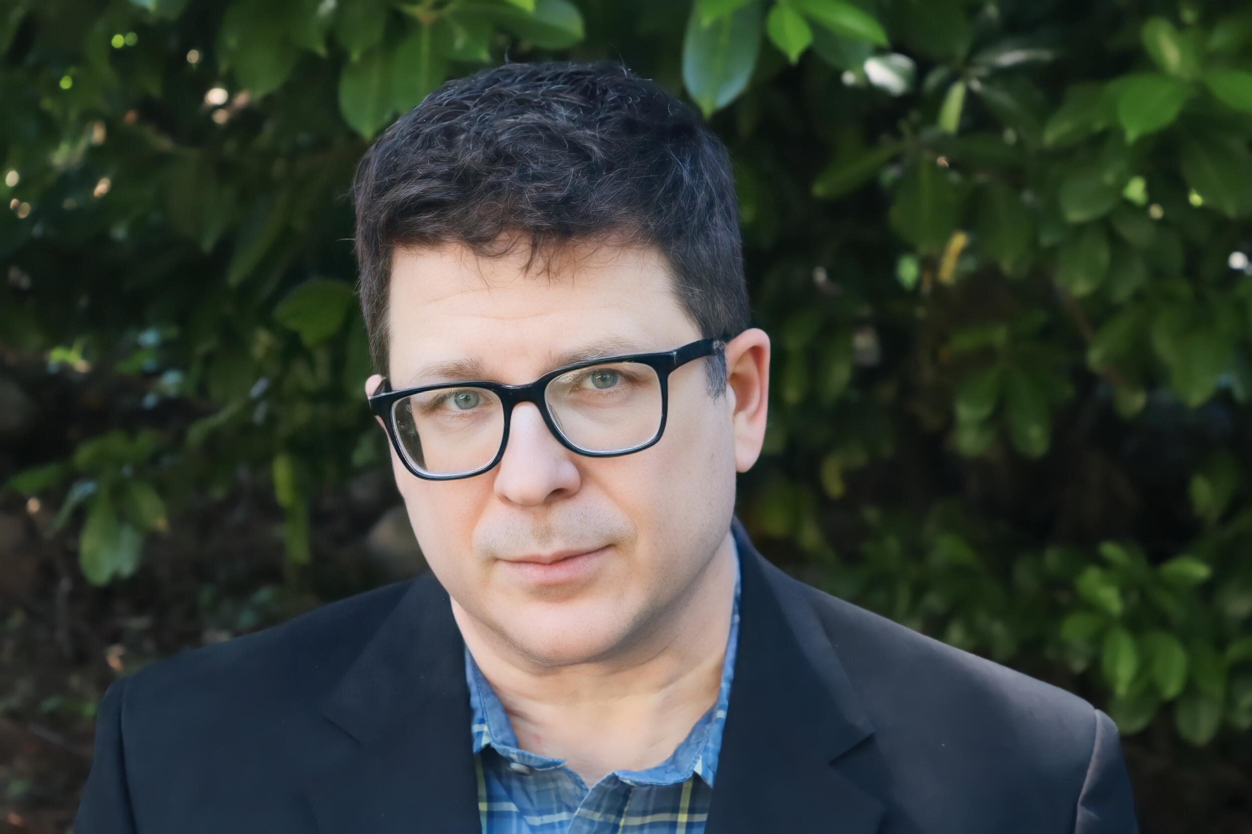 A photo of a man from the shoulders up. He is wearing glasses. 