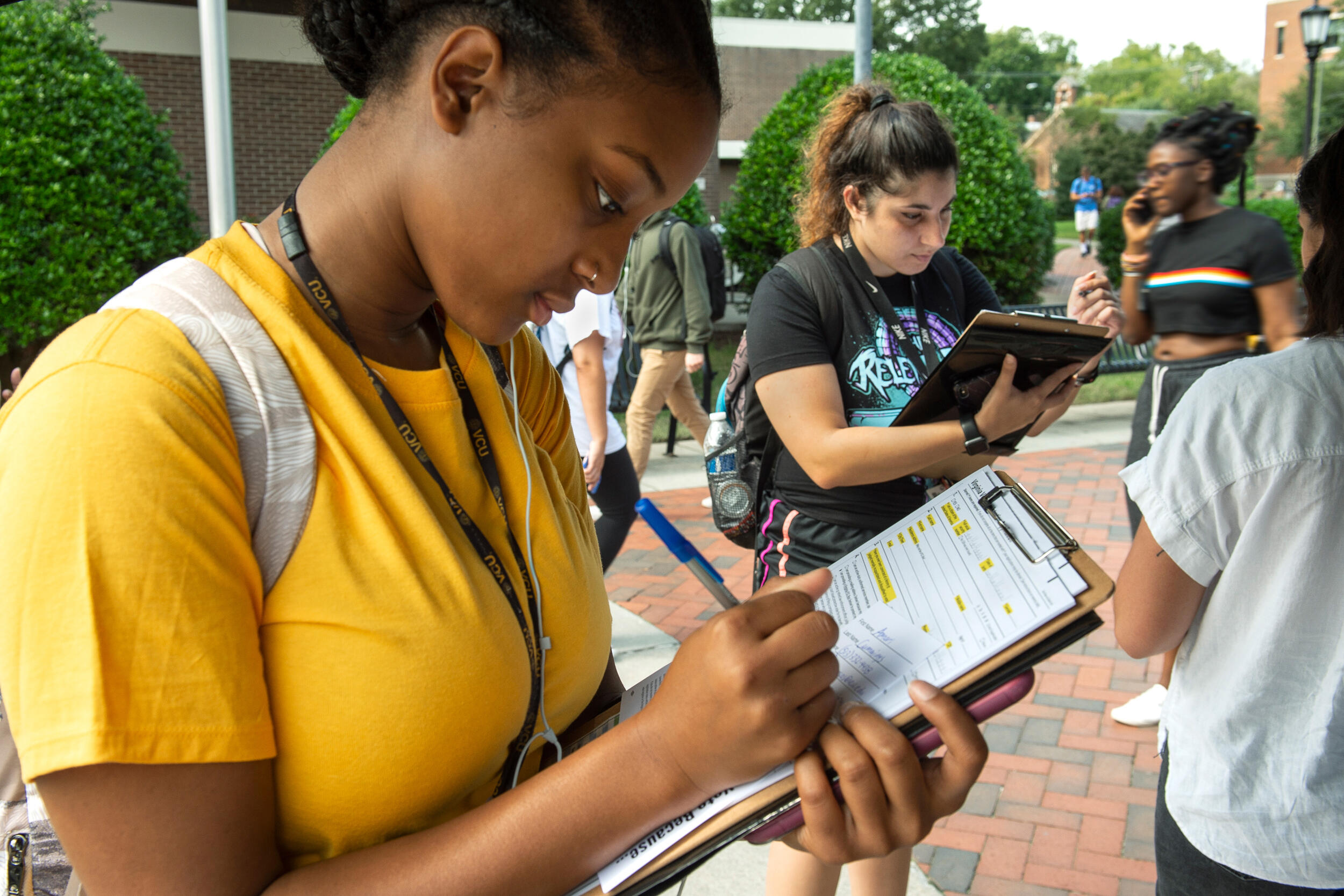 A student registers to vote on a clipboard while standing on a sidewalk with hedges in the background.