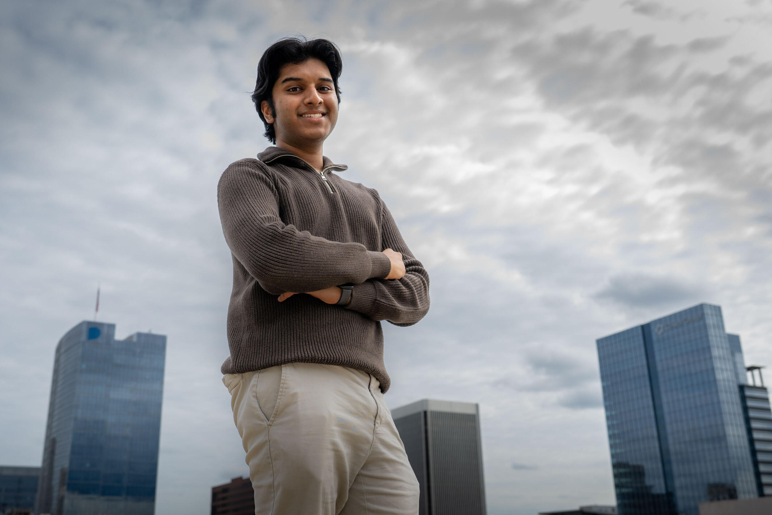 A photo of a man standing with his arms crossed against his chest while smiling. Behind him are skyscrapers.
