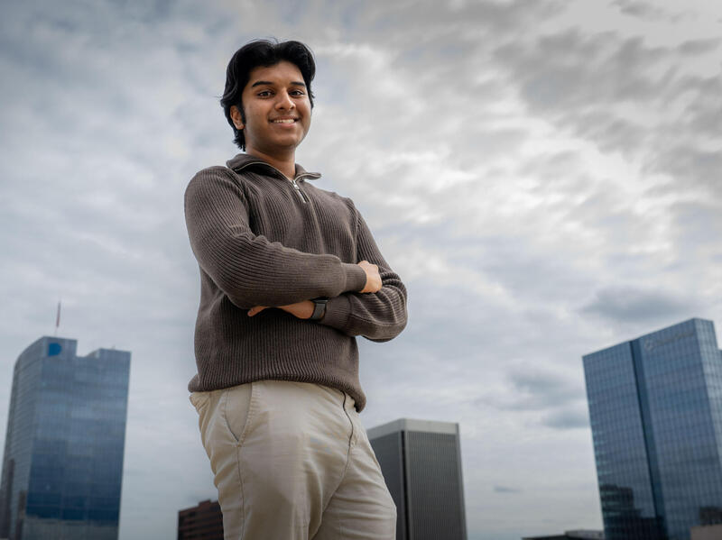A photo of a man standing with his arms crossed against his chest while smiling. Behind him are skyscrapers. 
