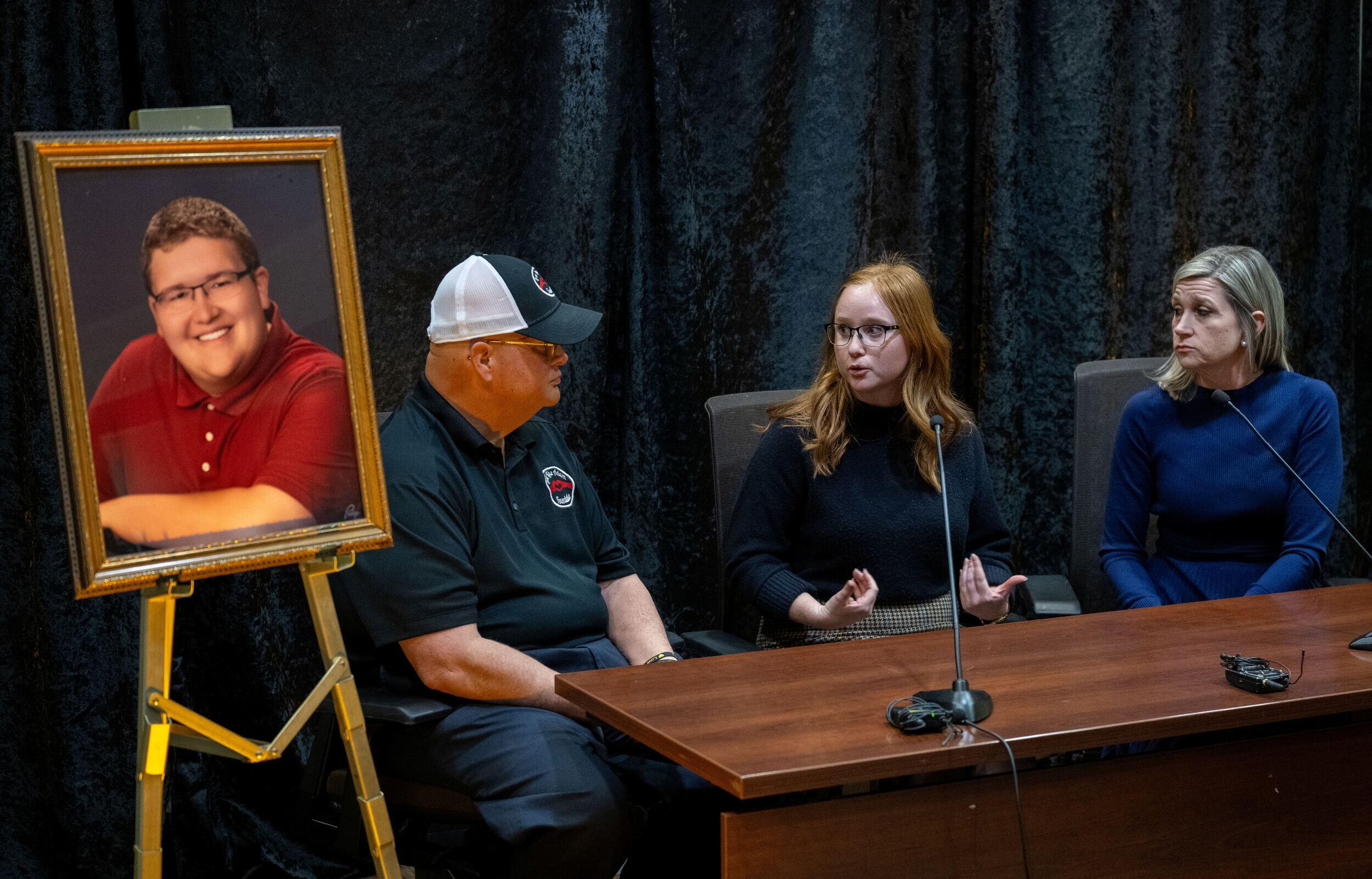 Three people sit at a desk speaking together with a microphone in front of them. To their right is a portrait on an easel of a young man in glasses and a red shirt.