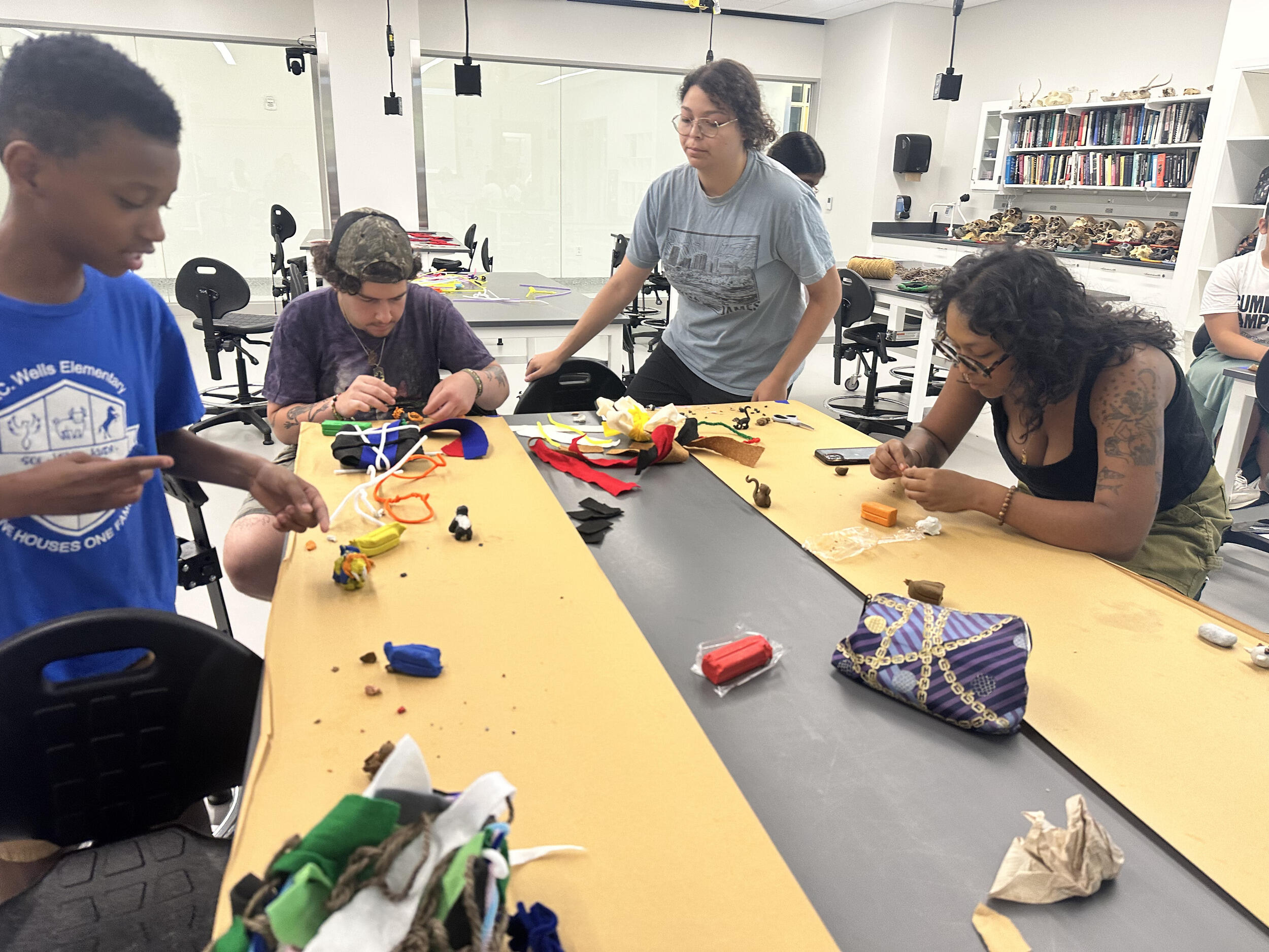 A photo of four people siting and standing around a table. There is clay people are using to create objects. 