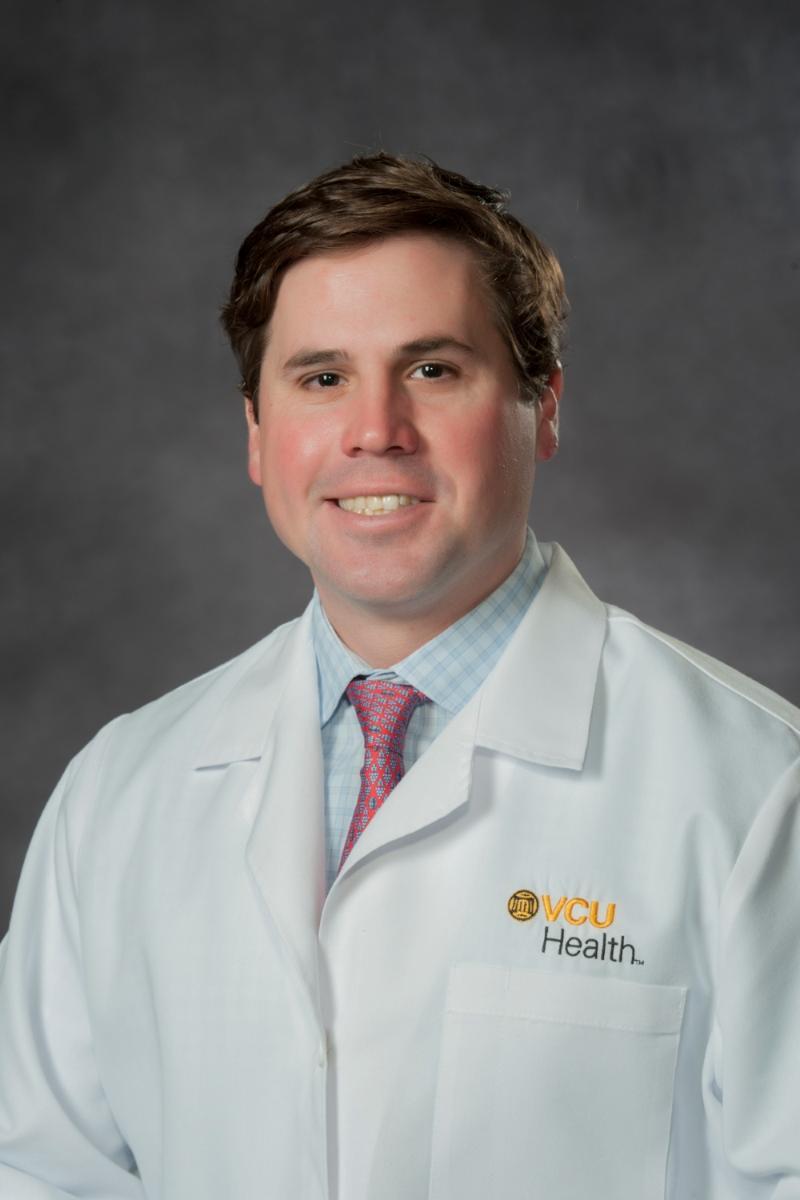 A photo of a man from the chest up. He is wearing a white doctor's coat that says \"VCU Health\" in black and yellow letters. 