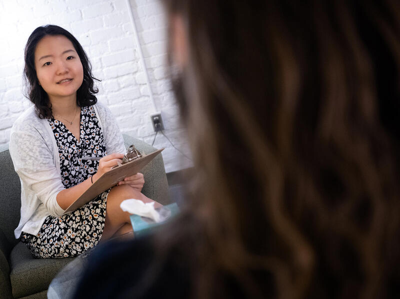 A photo of a woman holding a clipboard sitting on a chair talking to a person across from her who's face is not visable. 