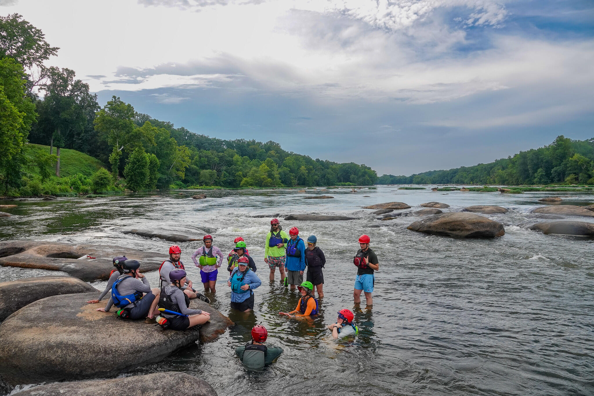 A photo of a group of people in the middle of a river. One man is standing in the middle of the group speaking. There is a circle of people around him listening. Some people are standing, and others are sitting on rocks or in the water. 