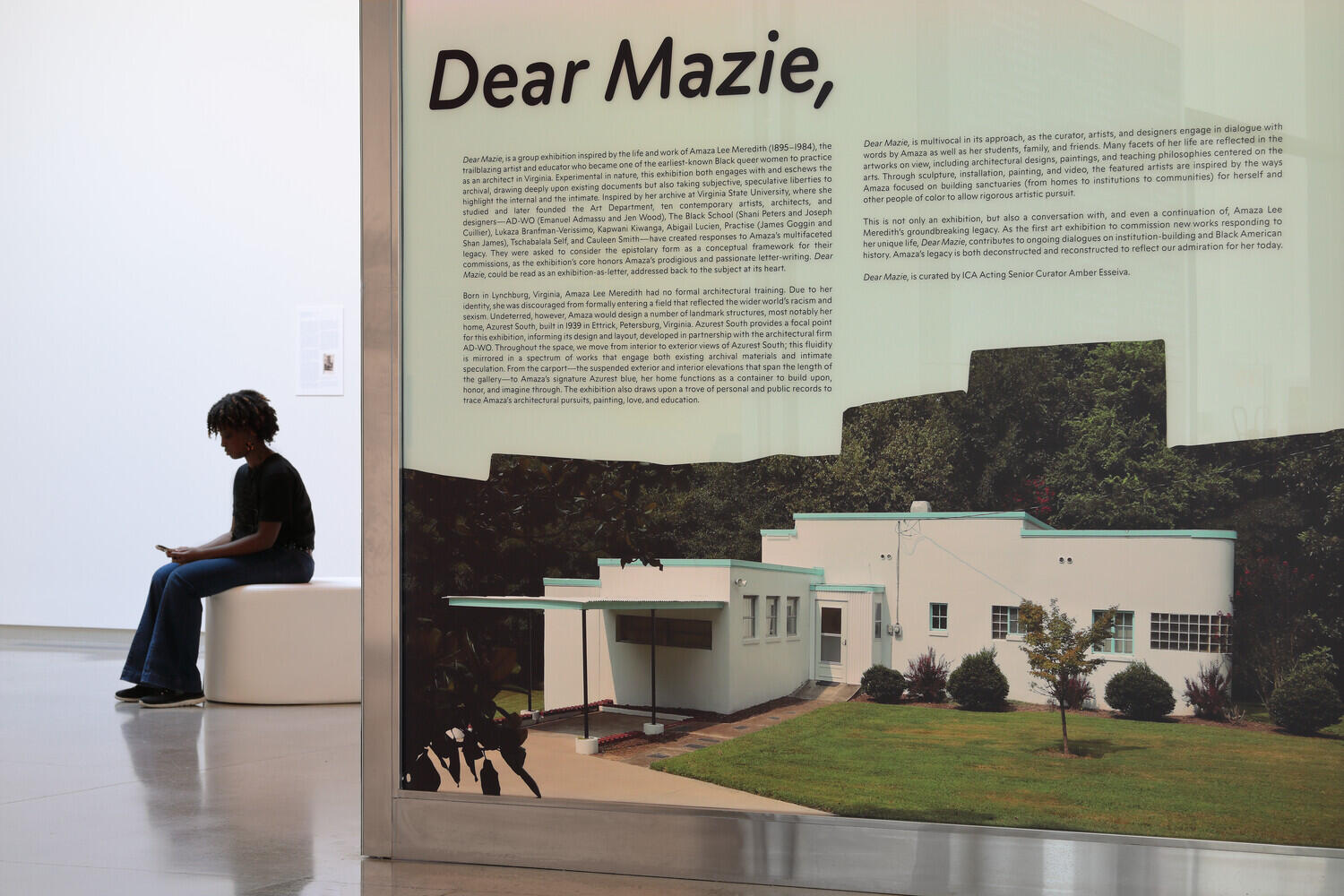 A photo of a sign that says \"Dear Mazie,\" with smaller text underneath that is illegible. There is a photo of a house underneath the text. To the left of the sign is a person sitting on a white bench.