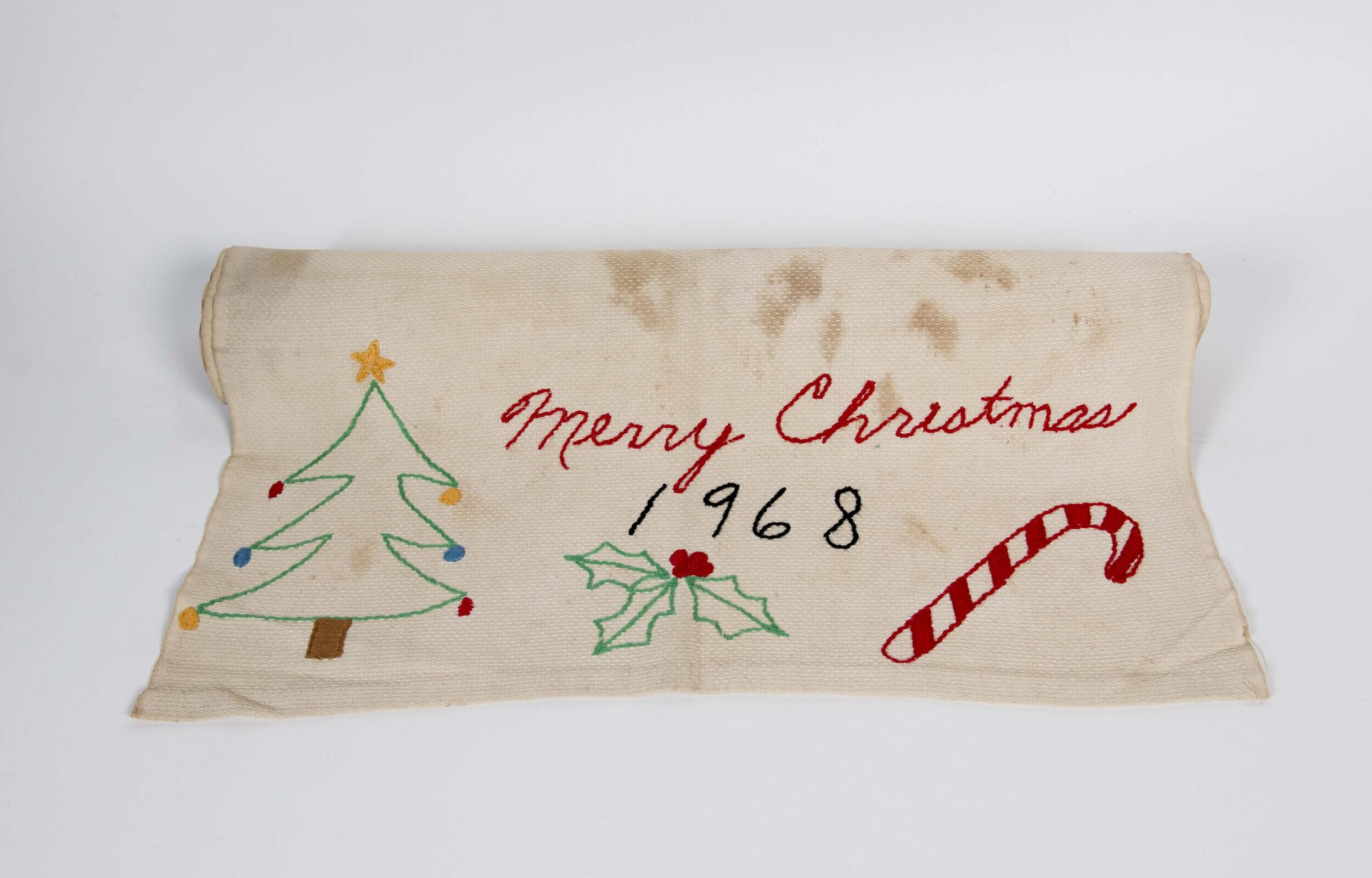 A photo of a piece of fabric that says \"Merry Christmas\" in red letters and \"1968\" in black letters. On the fabric are embroidered illustrations of a christmas tree, holly berries, and a candy canes.