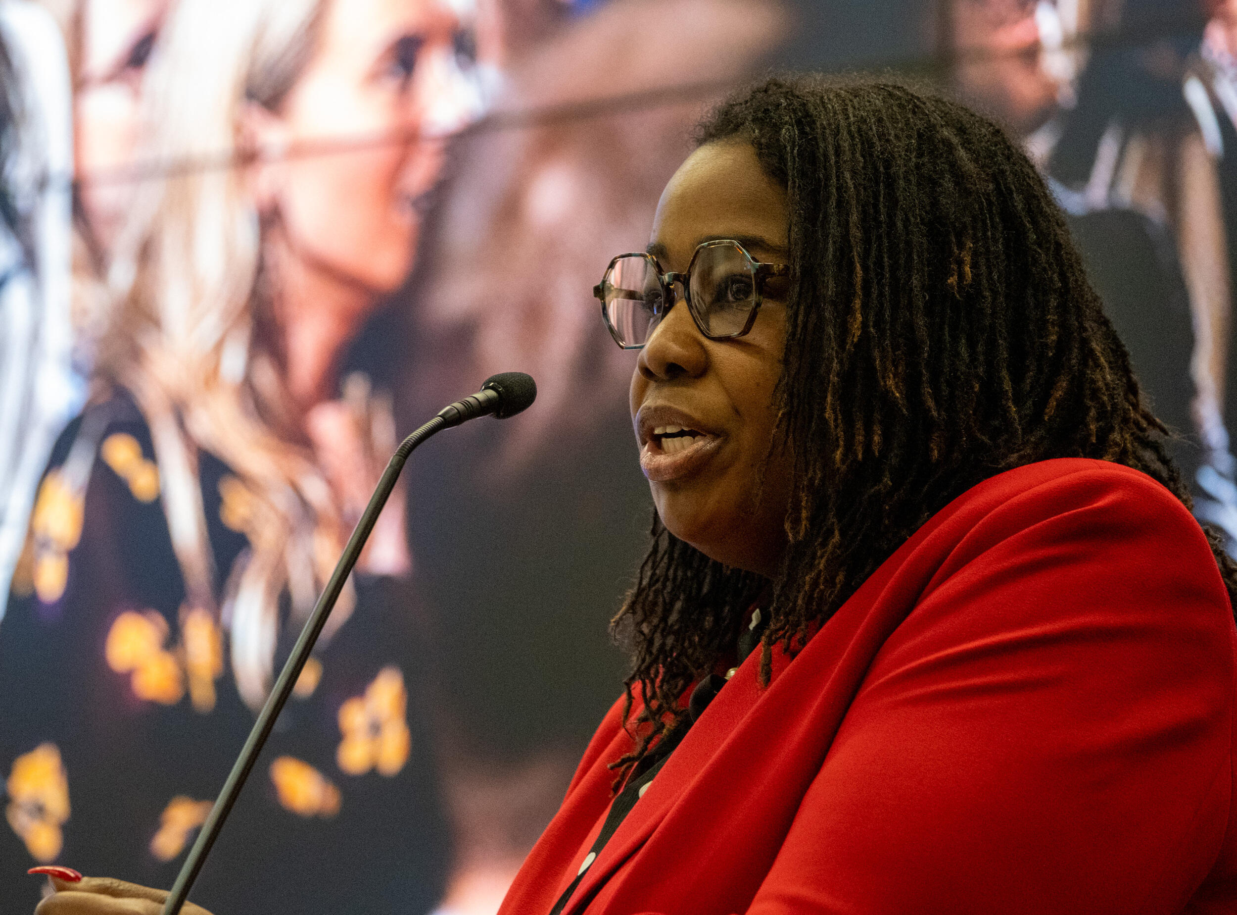 A photo of a woman from the chest up. She is facing the left and speaking into a microphone. 