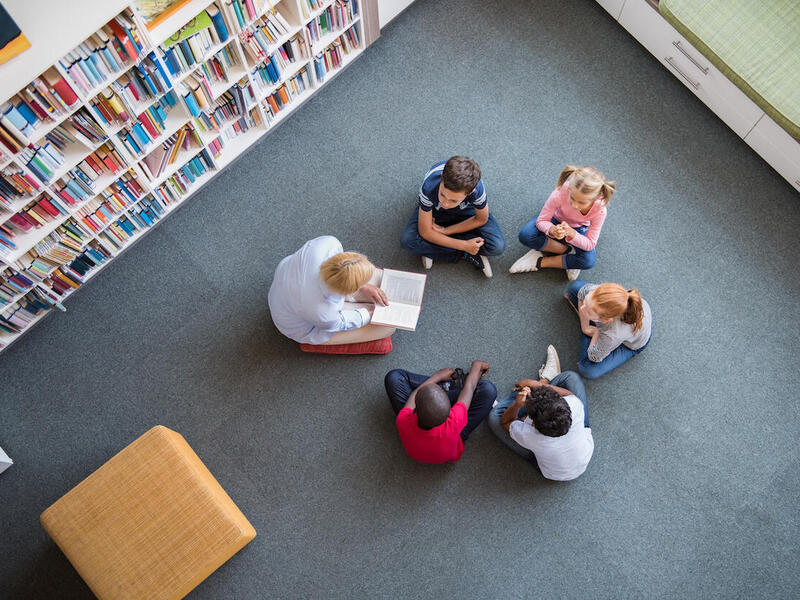 Students and teacher sit on the floor while teacher reads a book. 
