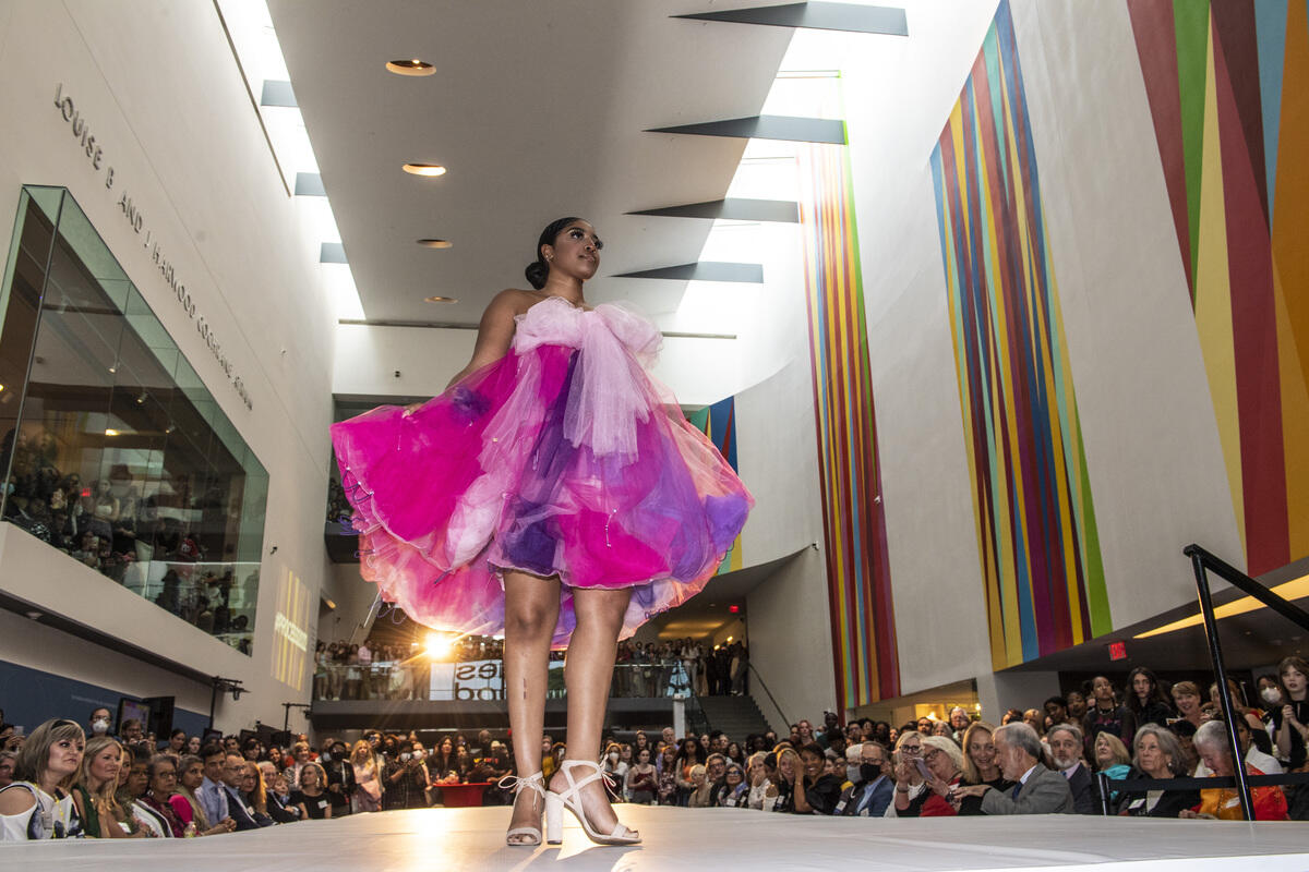 A fashion model wearing a pink and purple dress with white sandals walking a fashion runway surrounded by people at vmfa