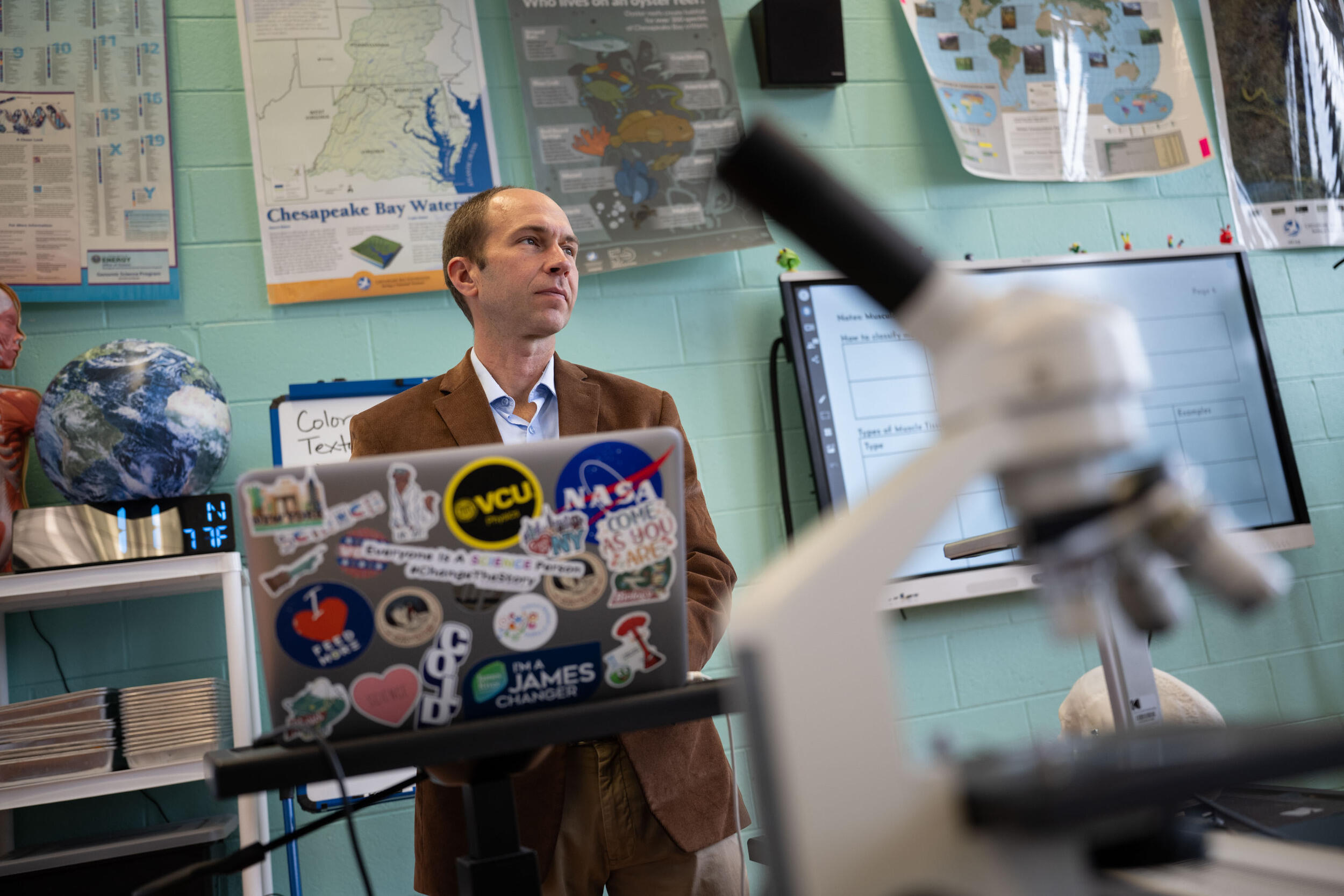 A photo of a man standing at the front of a classroom. He has a laptop in front of him covered in stickers. Behind him, the wall is covered in posters. There is a microscope in the foreground of the photo.