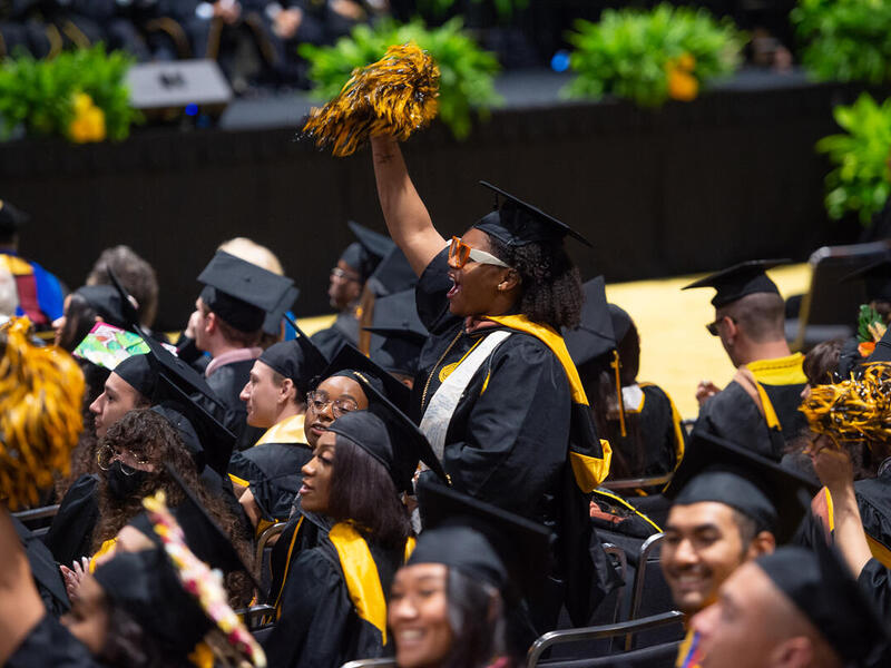 A group of people sitting in chairs wearing graduation caps and gowns. One woman stands in the middle of the crowd, cheering and waving a pompom in the air with her right arm. 