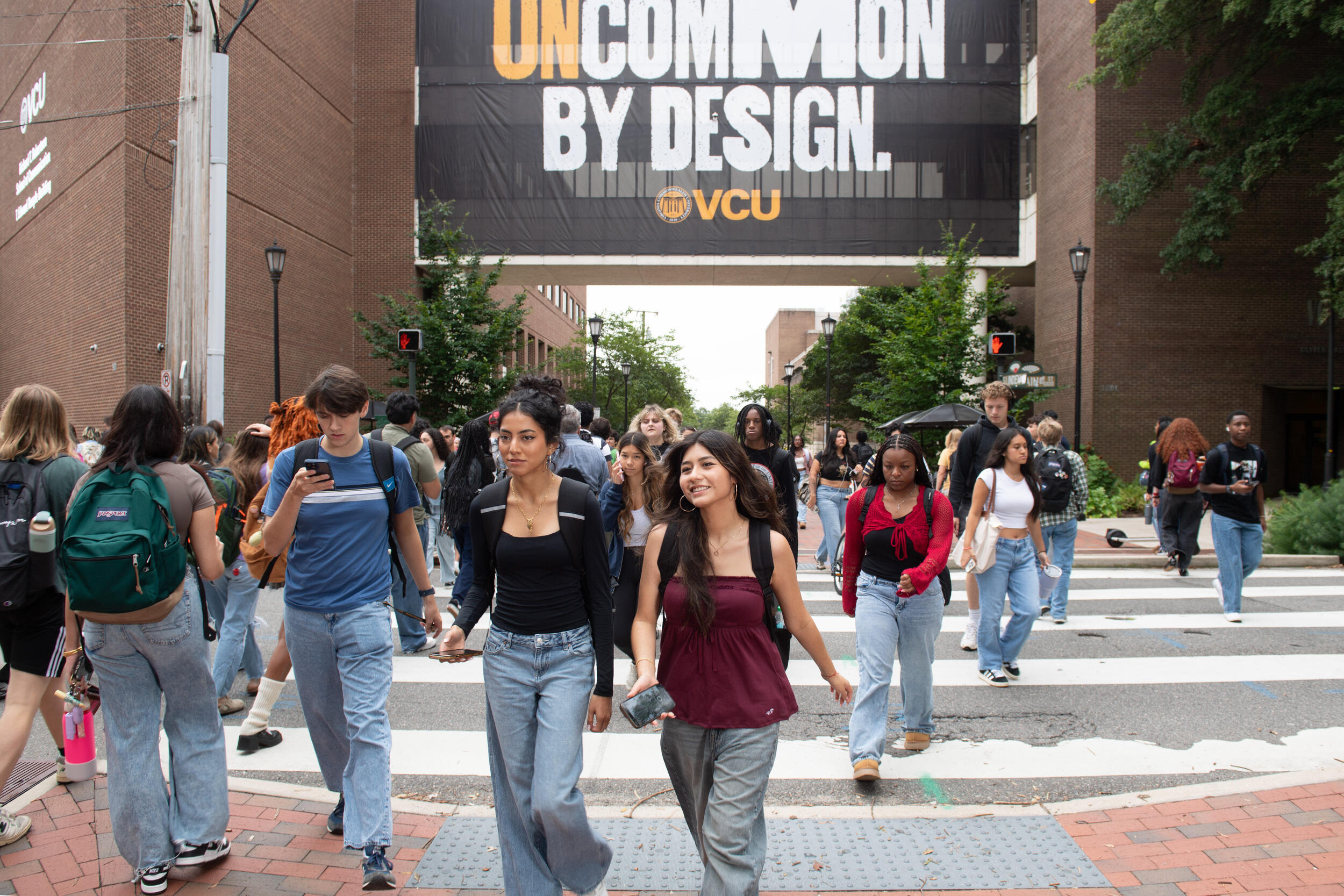 A photo of people walking on a sidewalk. Behind them is a sign that says \"UNCOMMON BY DESIGN VCU.\" 