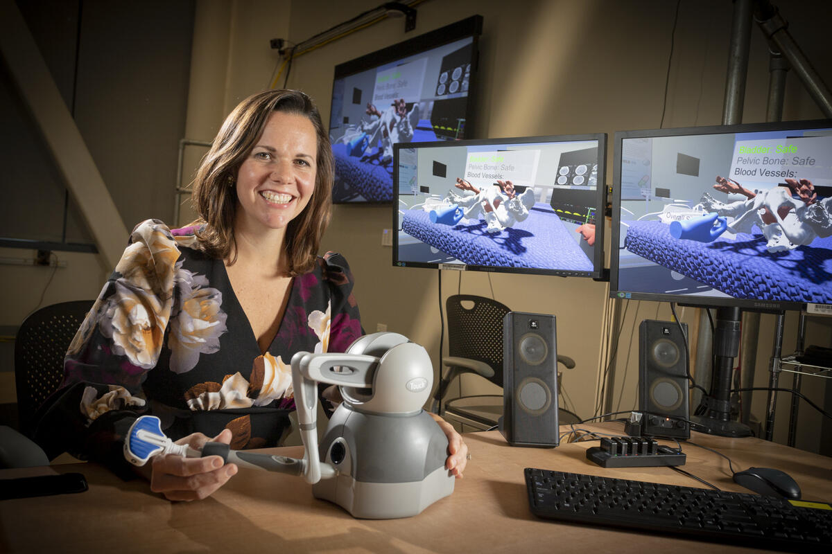 A woman smiling with her hands resting on a robotic arm. 