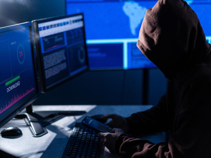 A photo of a hooded figure sitting at a desk with computer equipment to illustrate the PHP cybersecurity risks.