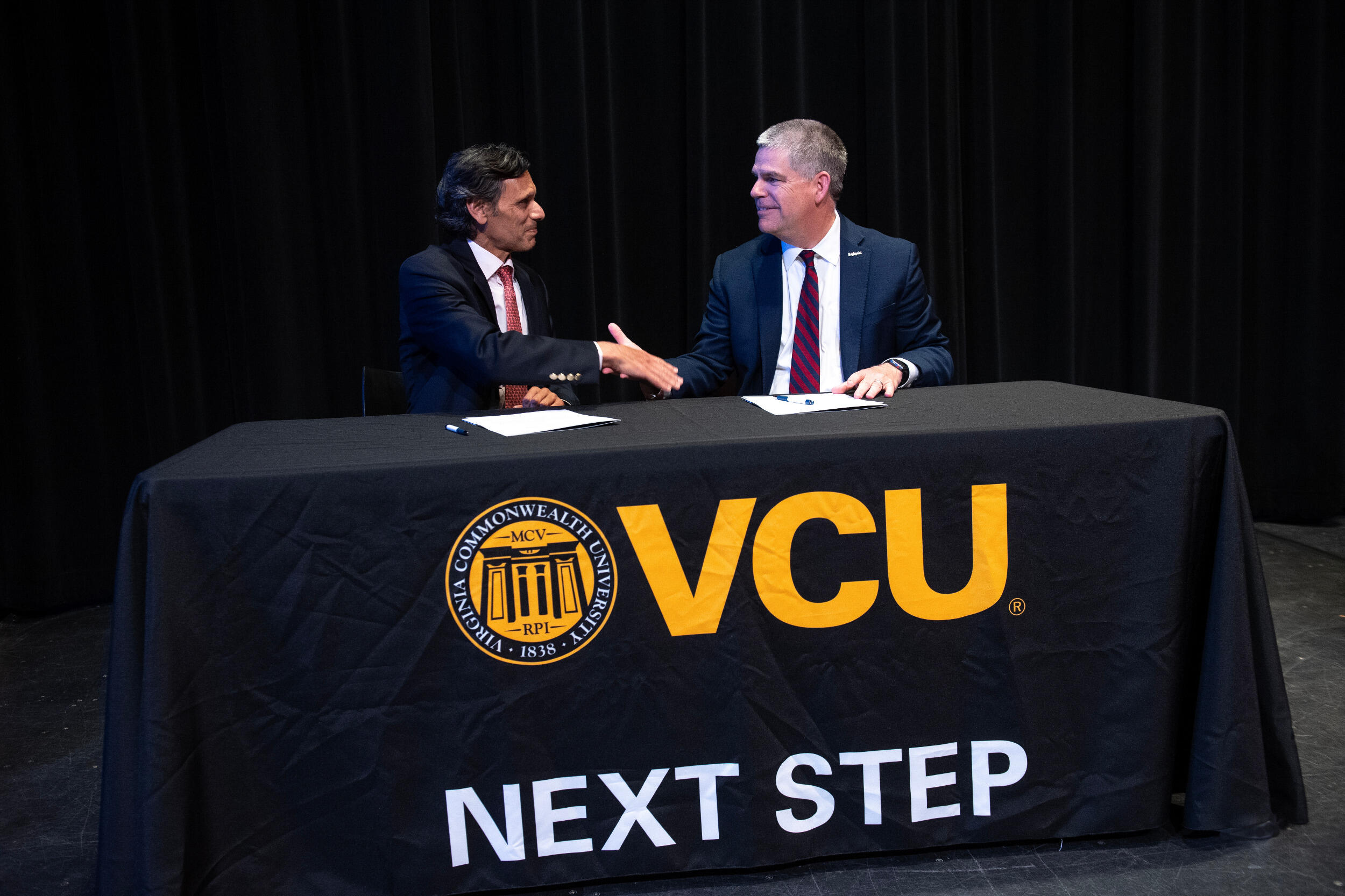 Two men in coats and ties sit at a table and shake hands.