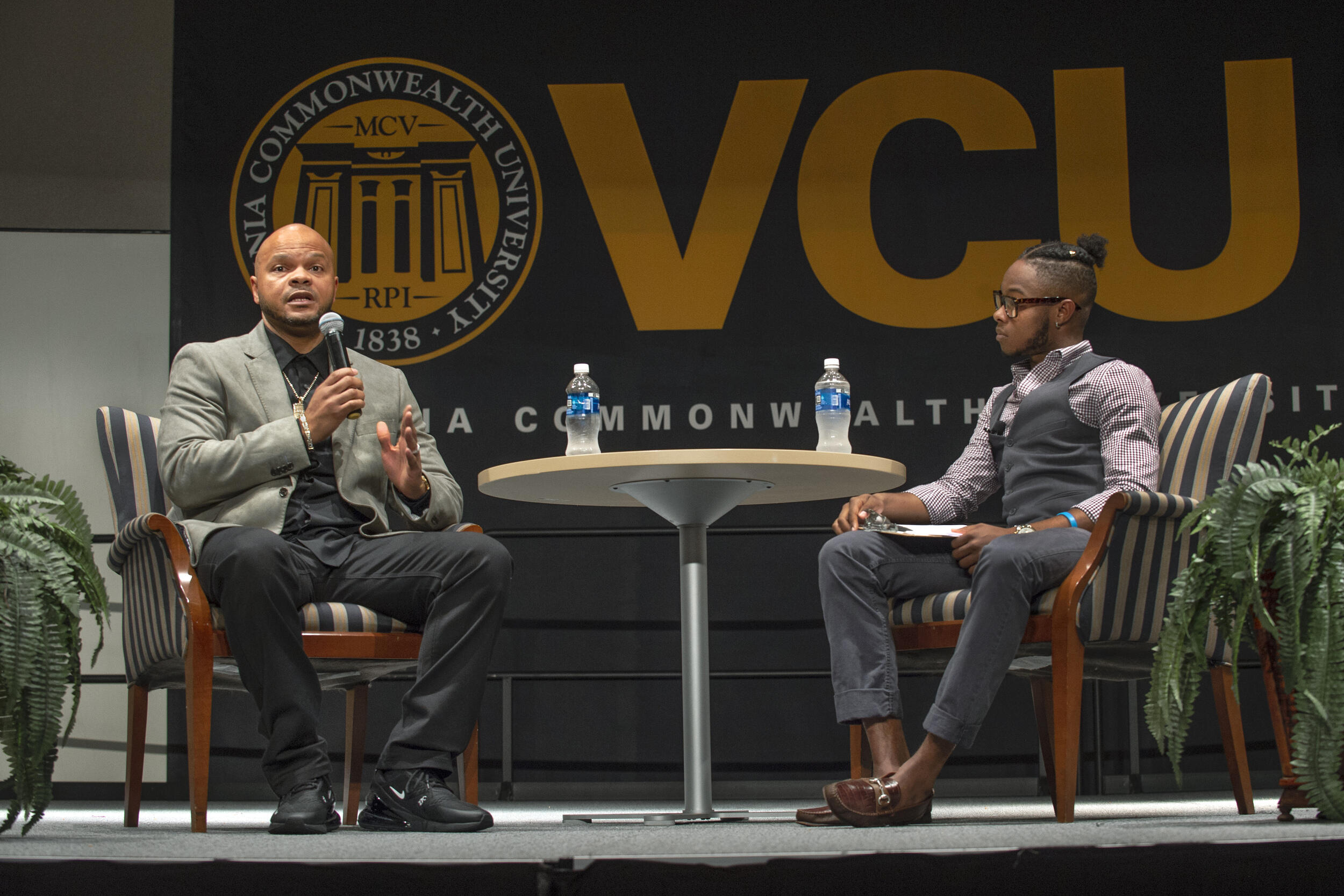 Kevin Richardson, left, addressing the audience. An event moderator sits in the seat on the right with a table is placed between the two chairs. The V C U logo is displayed behind the two people.