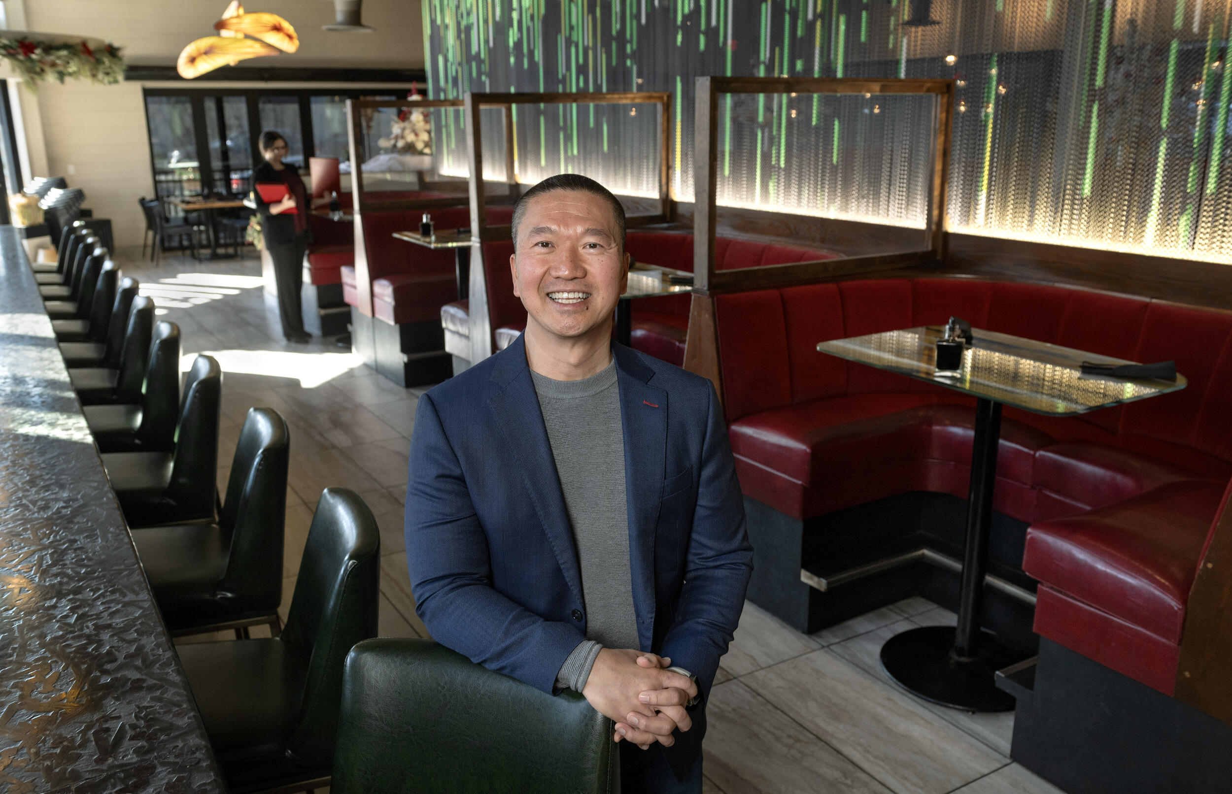 A photo of a man standing in a restaurant. He is leaning on one of the chairs along a bar. To his right are for red restaurant booths.