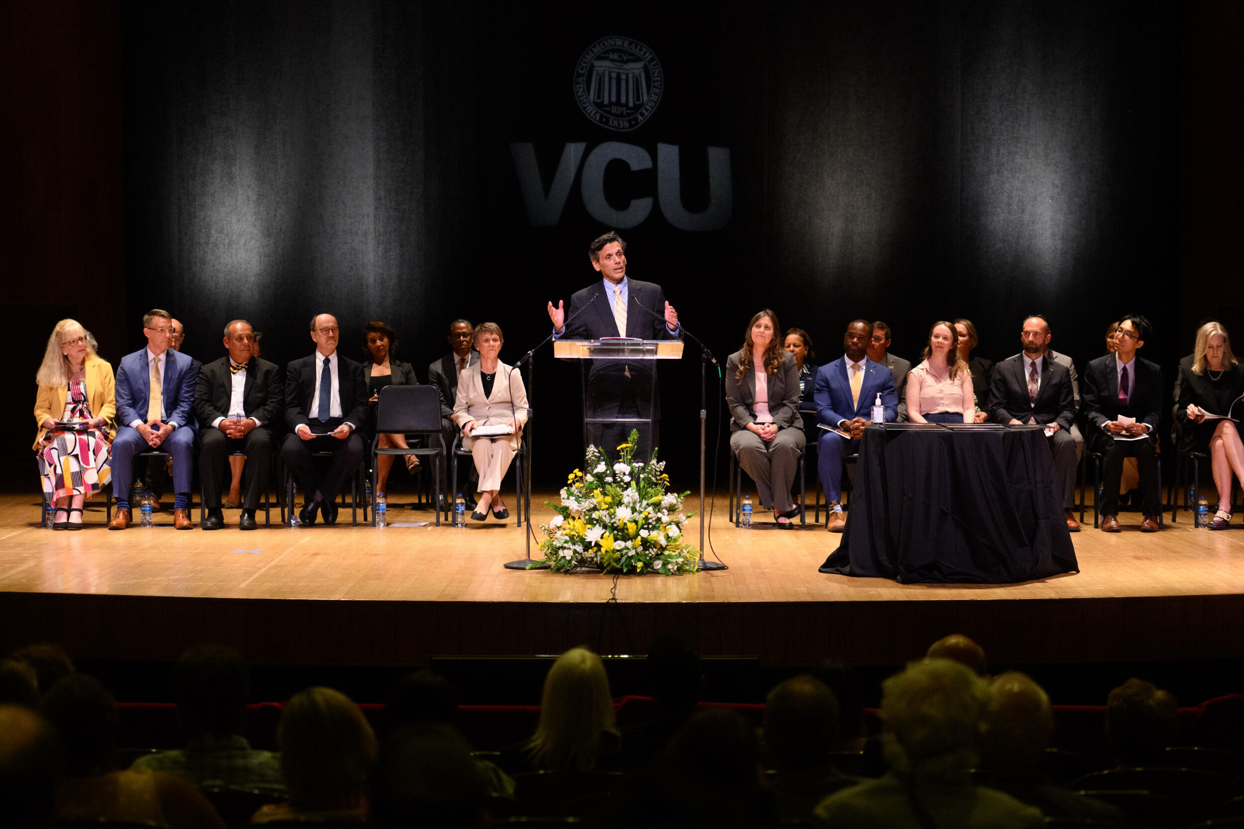 A man stands at a lectern on a stage with a group of people seated on chairs behind him.