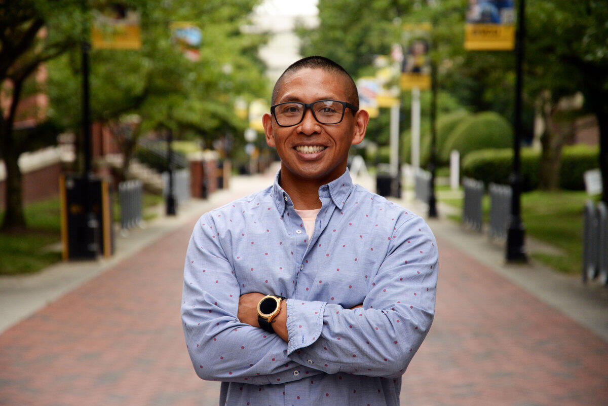 A photo of a man from the waist up with his arms crossed against his chest. He is standing in the middle of a brick sidewalk with trees and light posts on either side. 