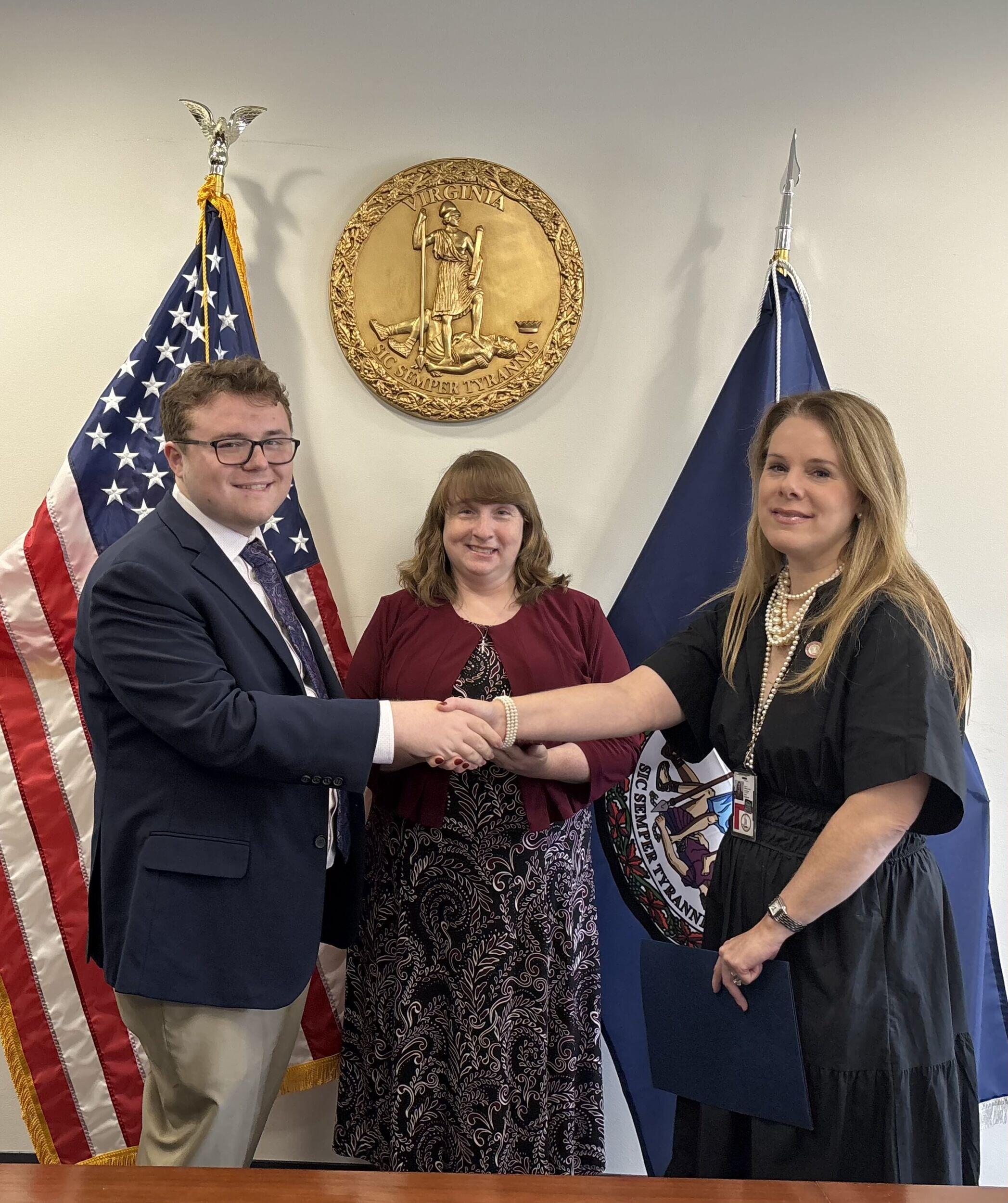 A photo of a man shaking hands with a woman in front of him. Behind the two people shaking hands is another woman. All three people are smiling and looking at the camera. 