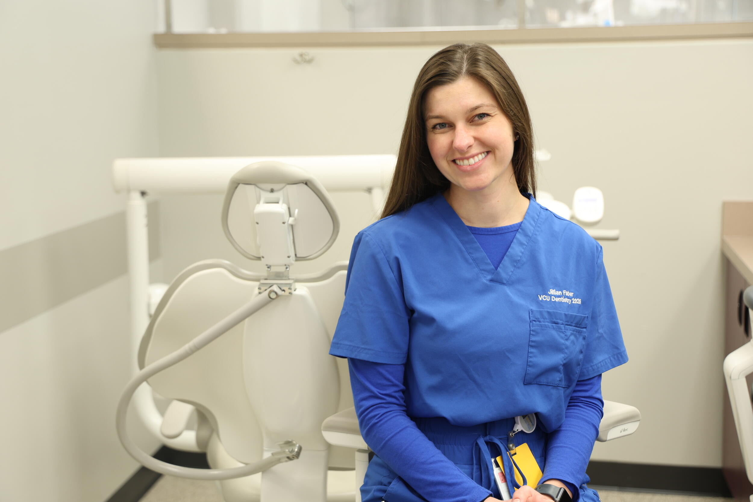 A photo of a woman wearing blue dental scrubs sitting next to a dental exam chair. 