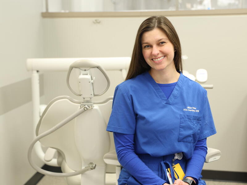 A photo of a woman wearing blue dental scrubs sitting next to a dental exam chair. 