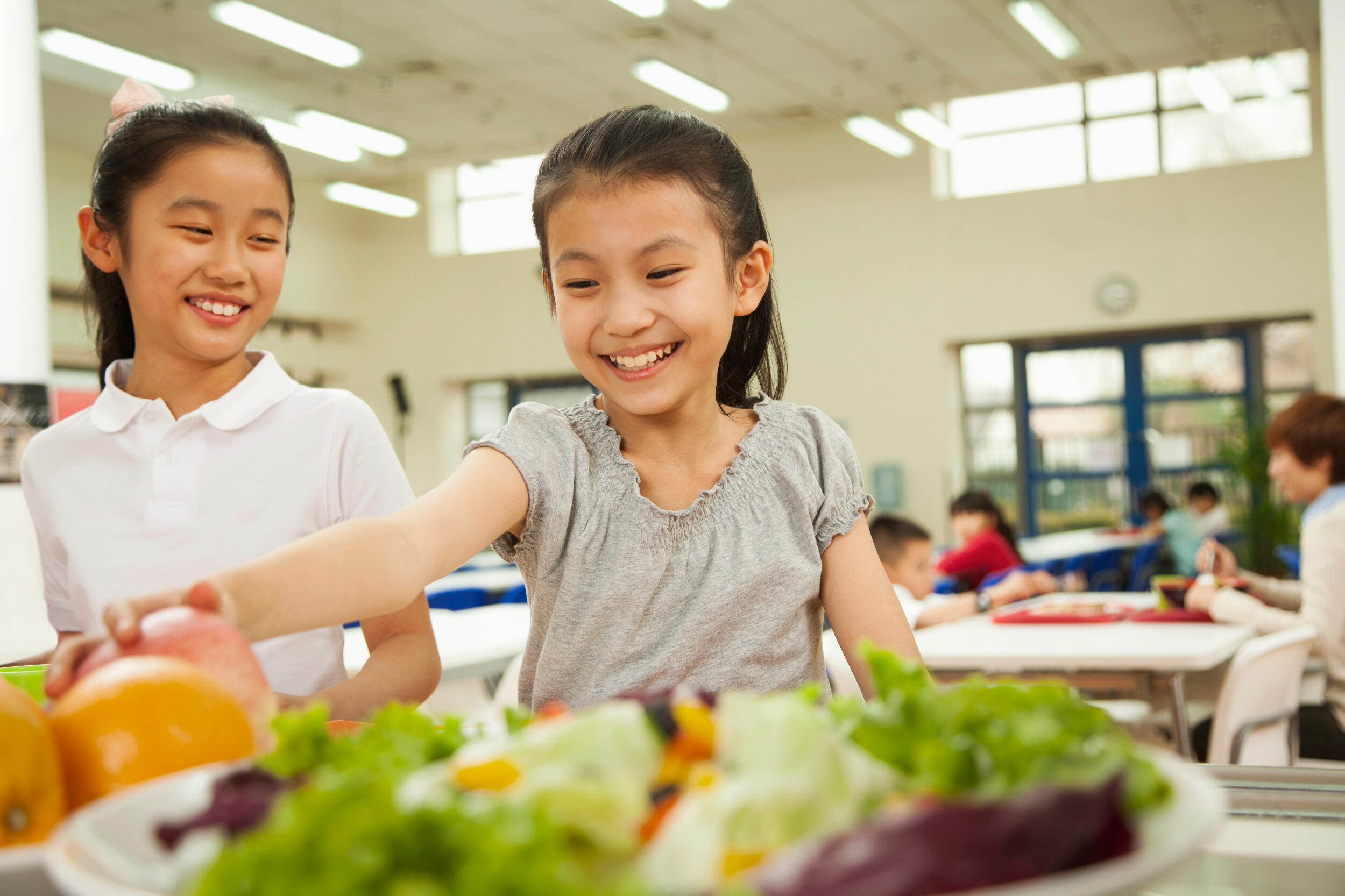 Two young girls smile and select food in a school cafeteria-type environment.