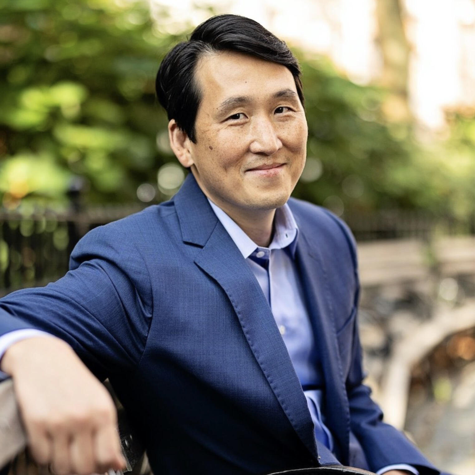A photo of a man from the waist up. He is sitting on a bench. 
