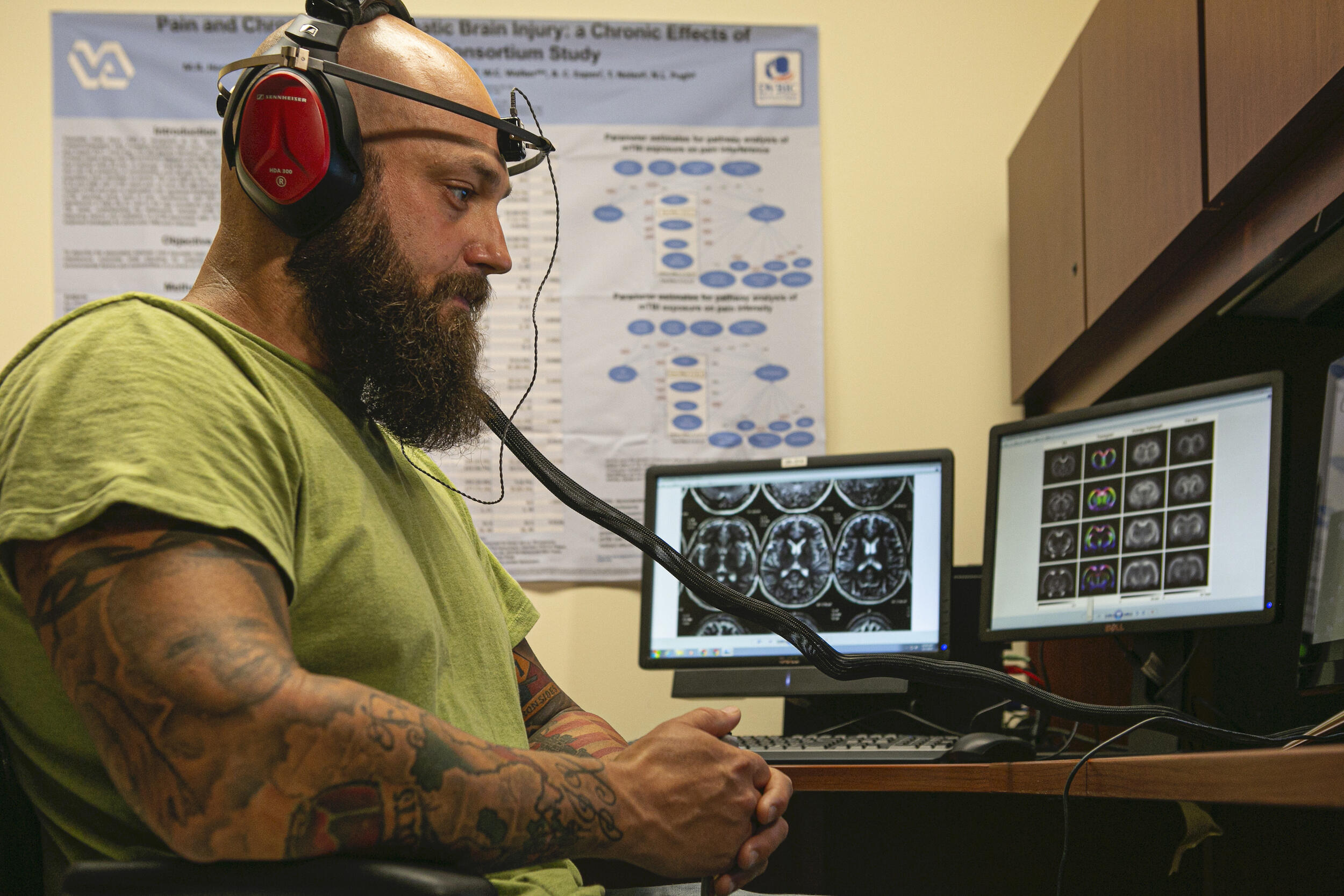 A person wearing headphones and a band around their head sits in a chair. Computer screens display images of brain scans.