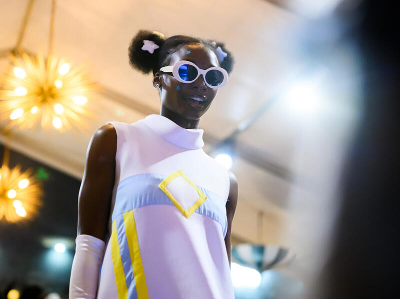 A photo of a woman from the waist up. She is walking and wearing a turtle neck dress, long gloves, and sunglasses. 