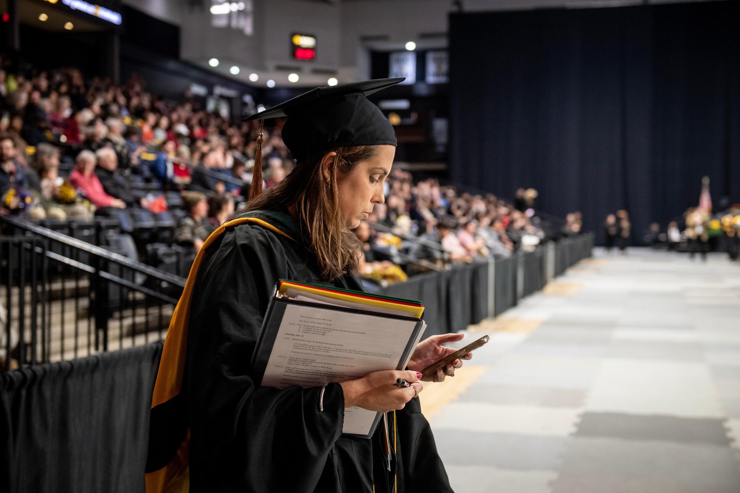 A photo of a woman wearing a graduation cap and gown holding a binder and a folder in one hand and looking at a cell phone in the other. Behind her is a crowd of people sitting in stadium seats. 