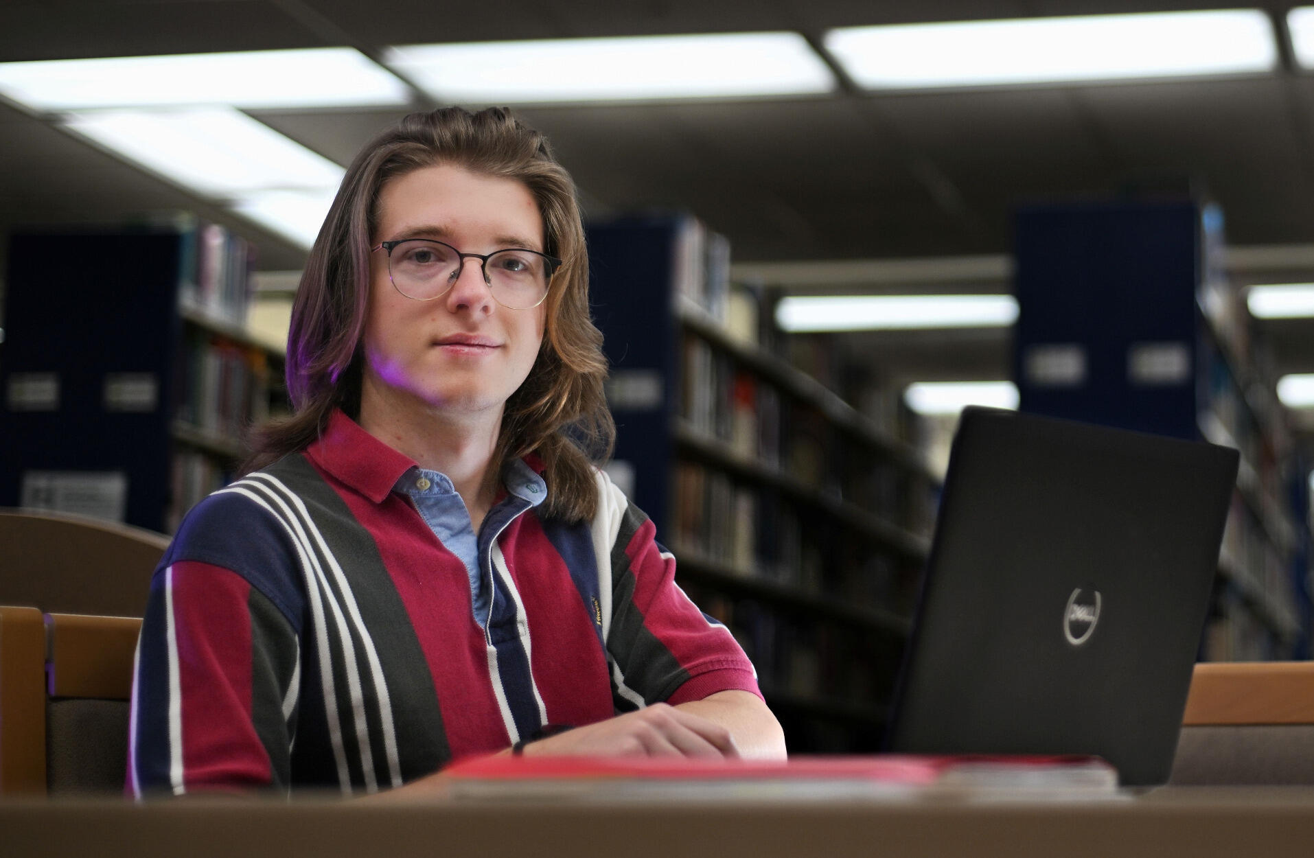 Man in glasses sitting at a table in a library with an open laptop.